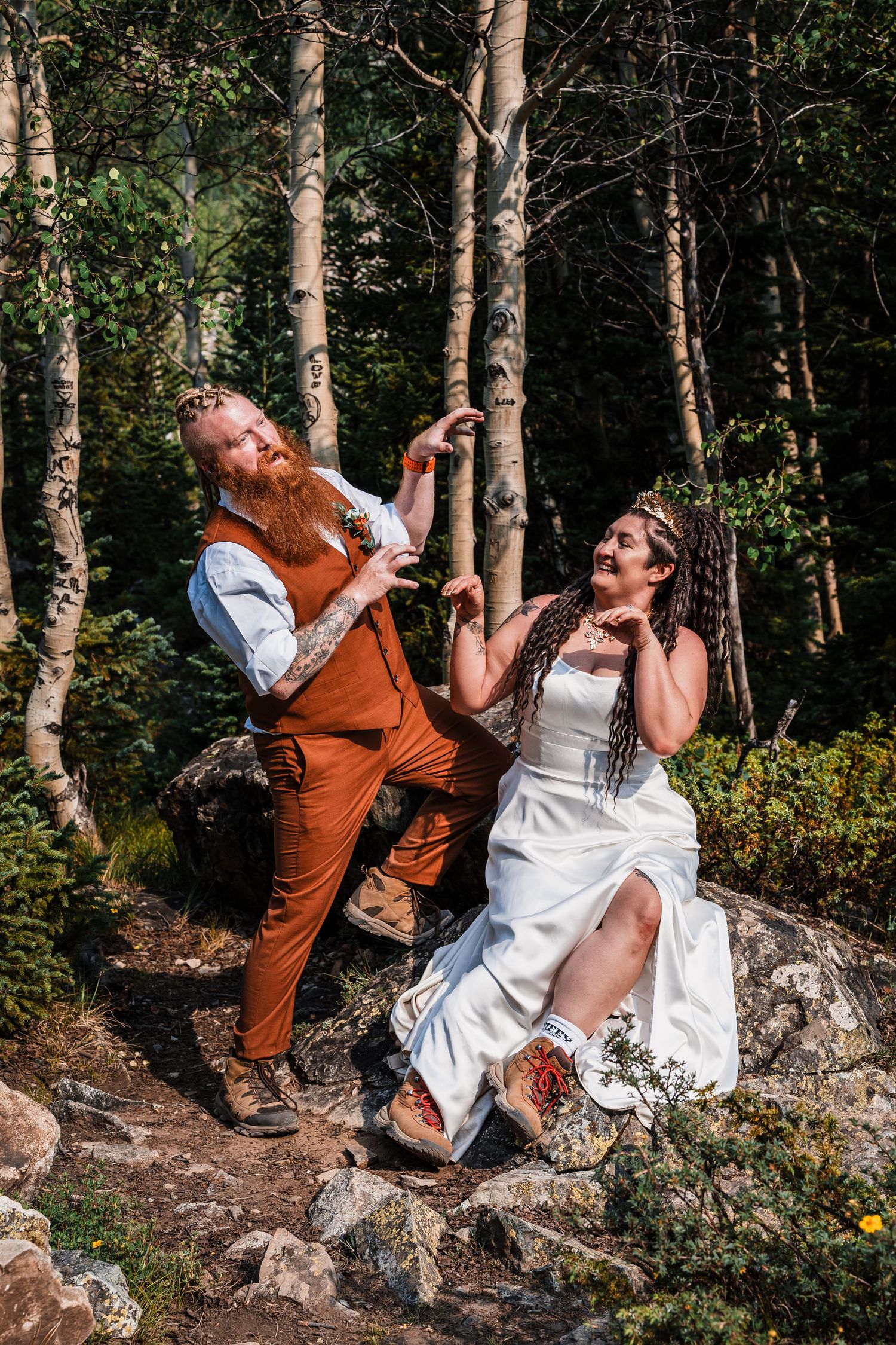 Laughing bride and groom posing playfully on rocks among aspen trees in forest wedding photos.