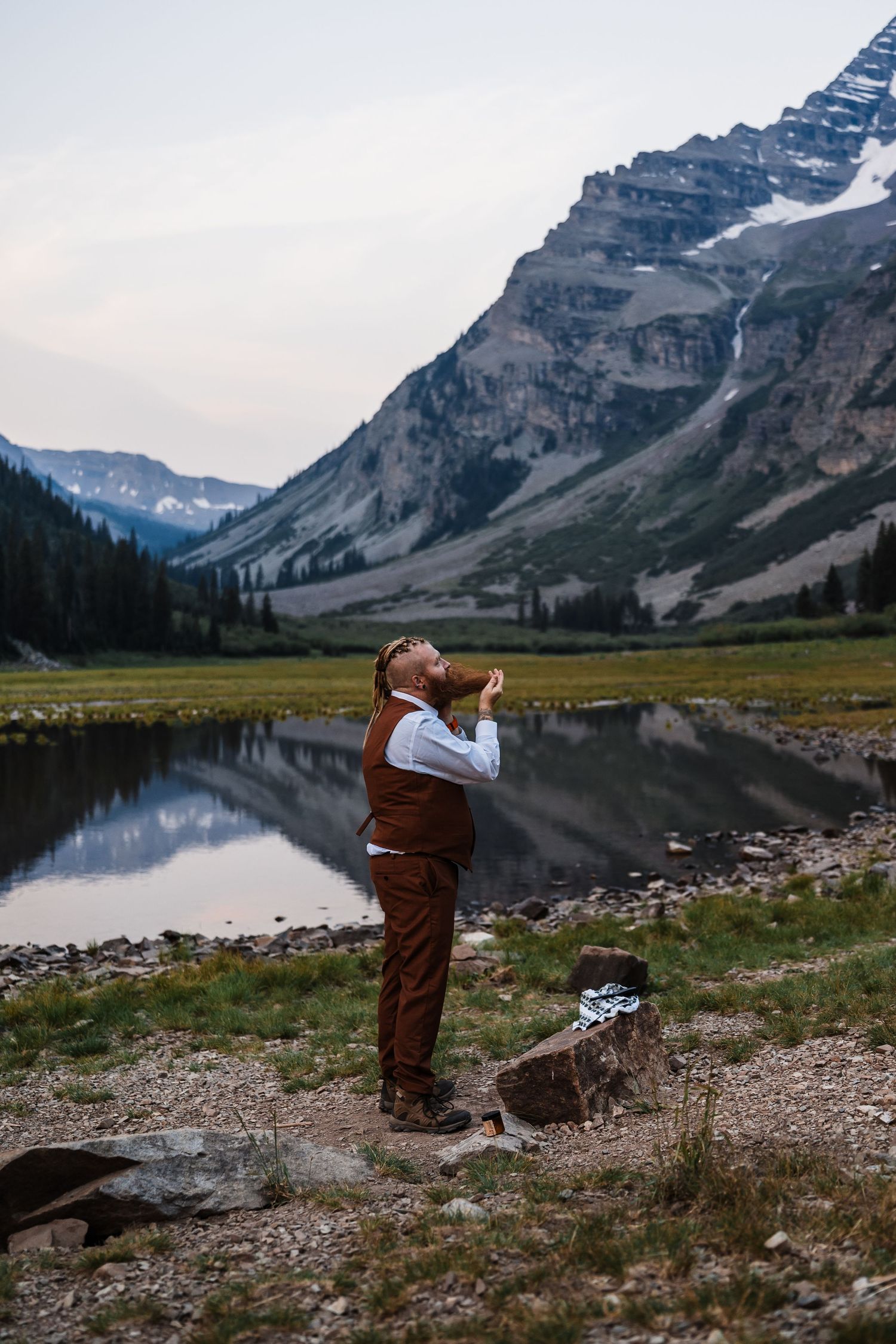 Groom in brown vest and pants drinking by a mountain lake with stunning rocky peak reflection.