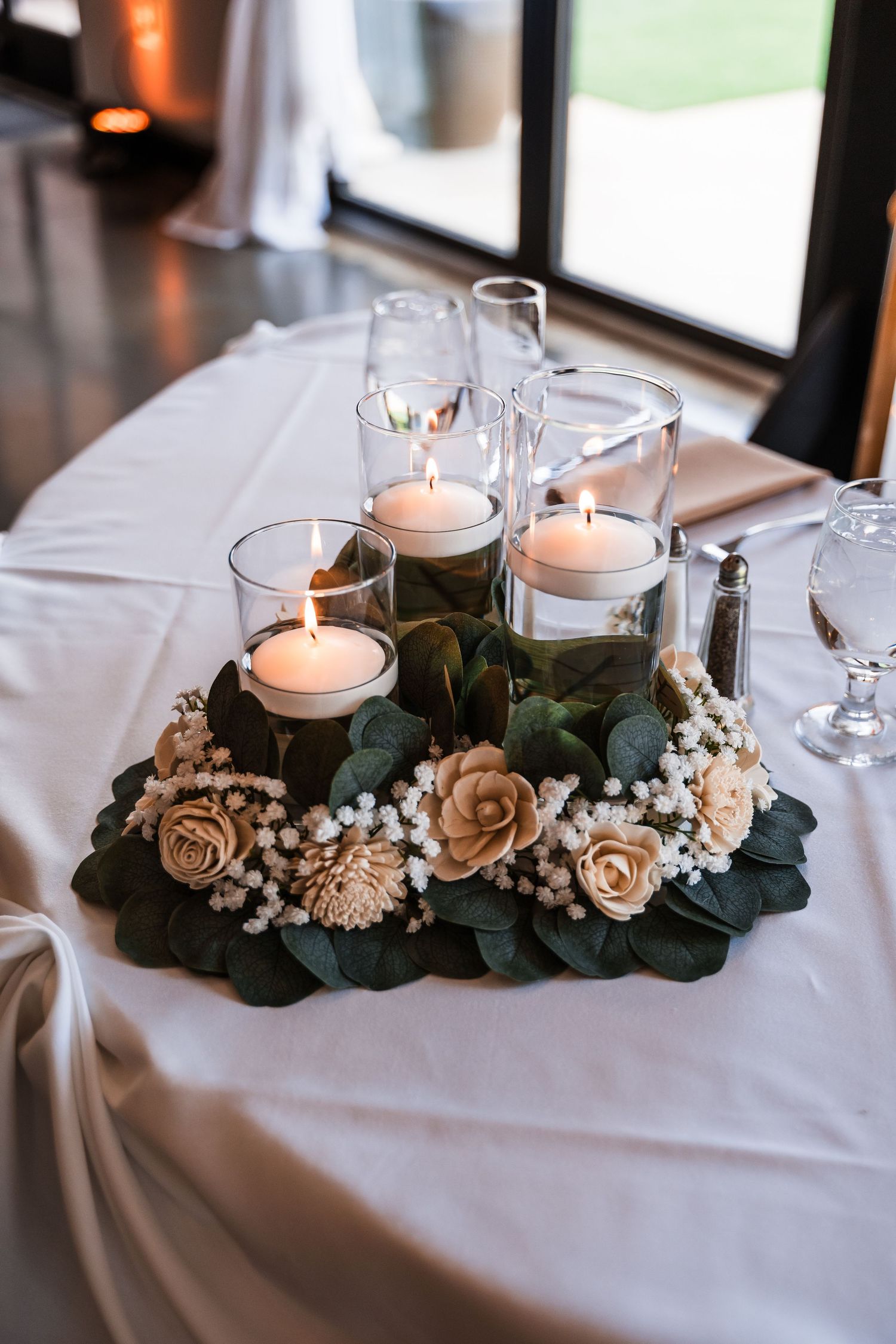 Elegant wedding centerpiece with lit candles in glass holders surrounded by flowers and greenery.
