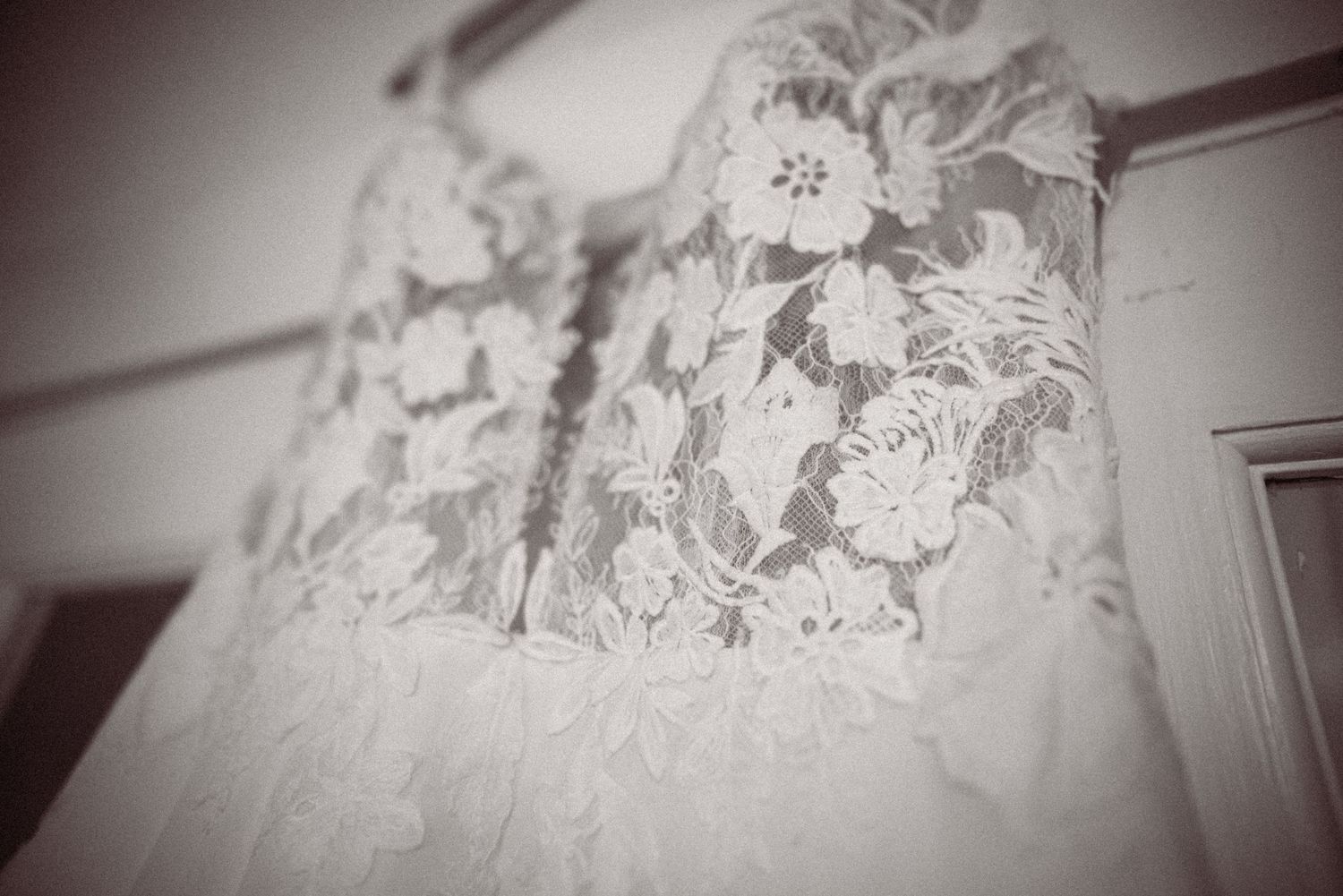 Elegant white lace wedding dress with floral embroidery hanging on display in a soft black and white photograph.