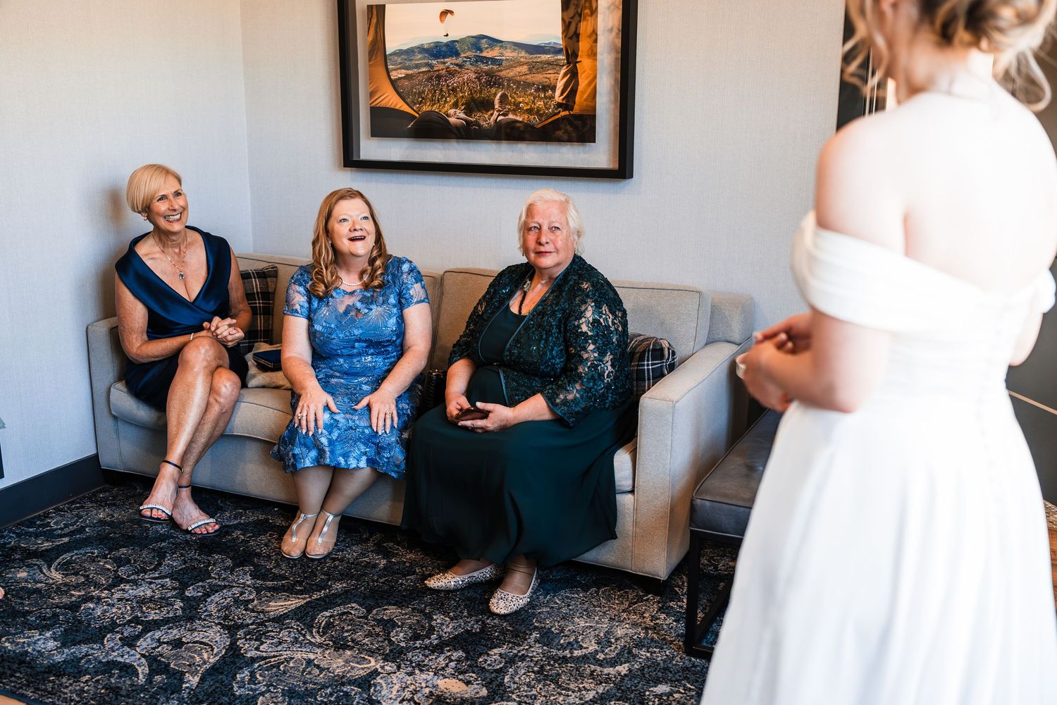 Three women smile and react joyfully while a bride in white dress reveals her wedding gown.