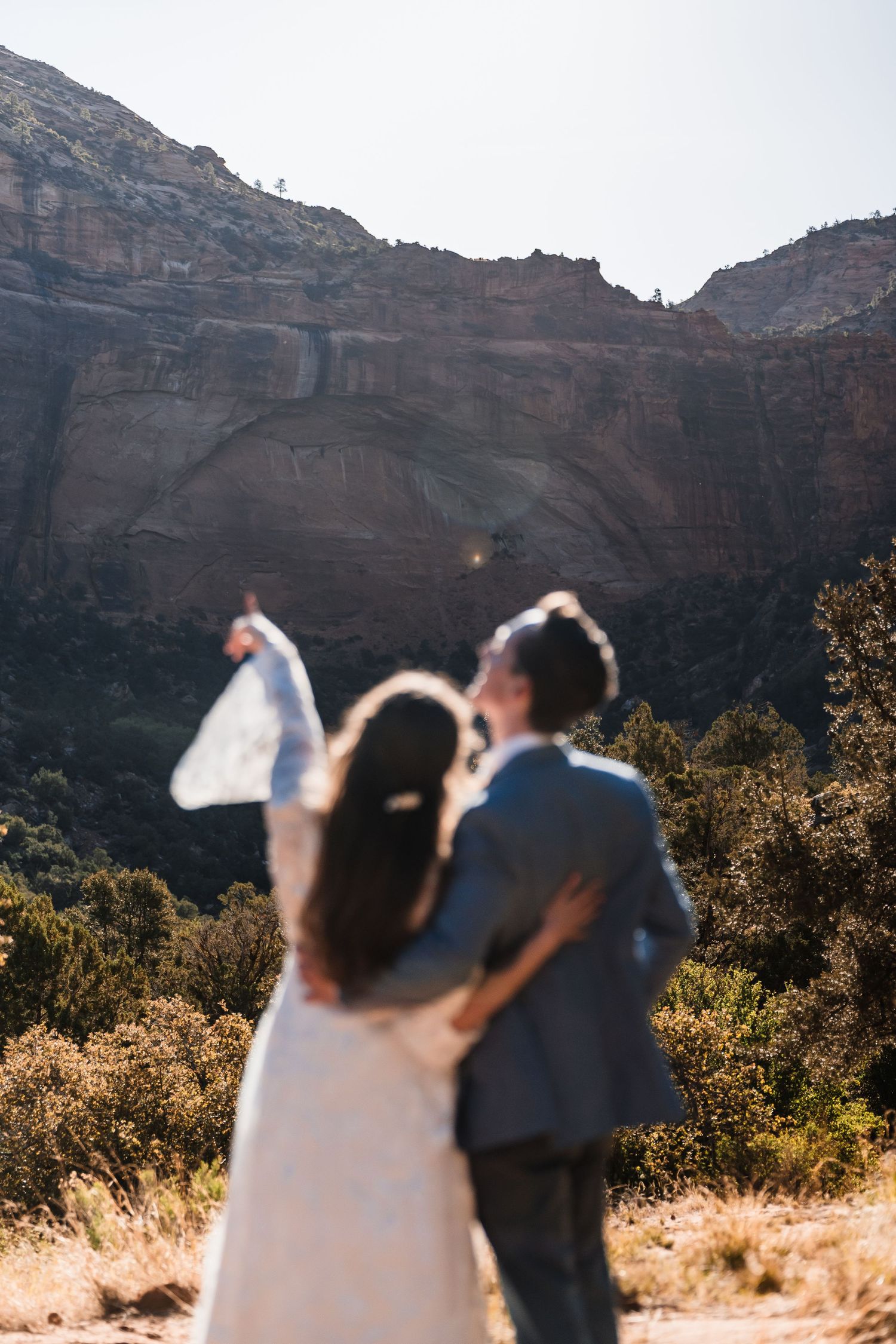 Bride and groom embrace while gazing at stunning red rock canyon cliffs during their outdoor desert wedding ceremony.