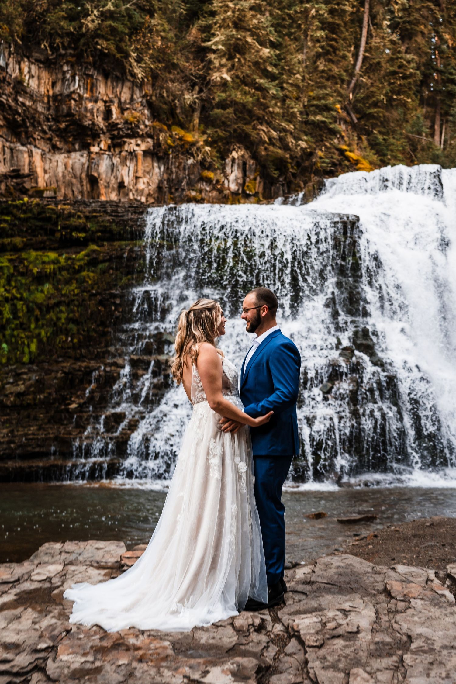 Scenic waterfall backdrop with couple in wedding attire on rocky outcrop.