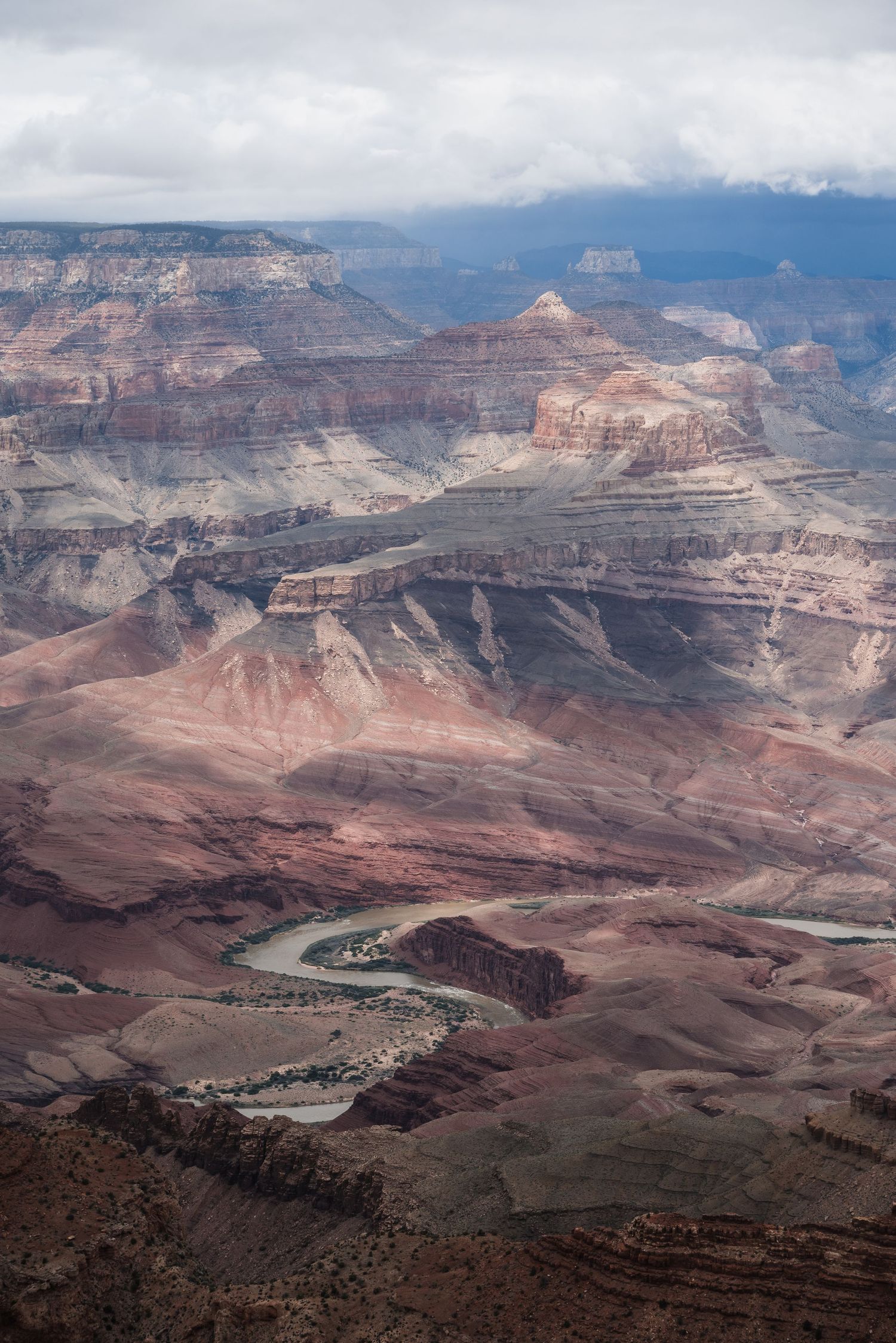 The Colorado River winds through the layered red rock formations of the Grand Canyon under dramatic cloudy skies.