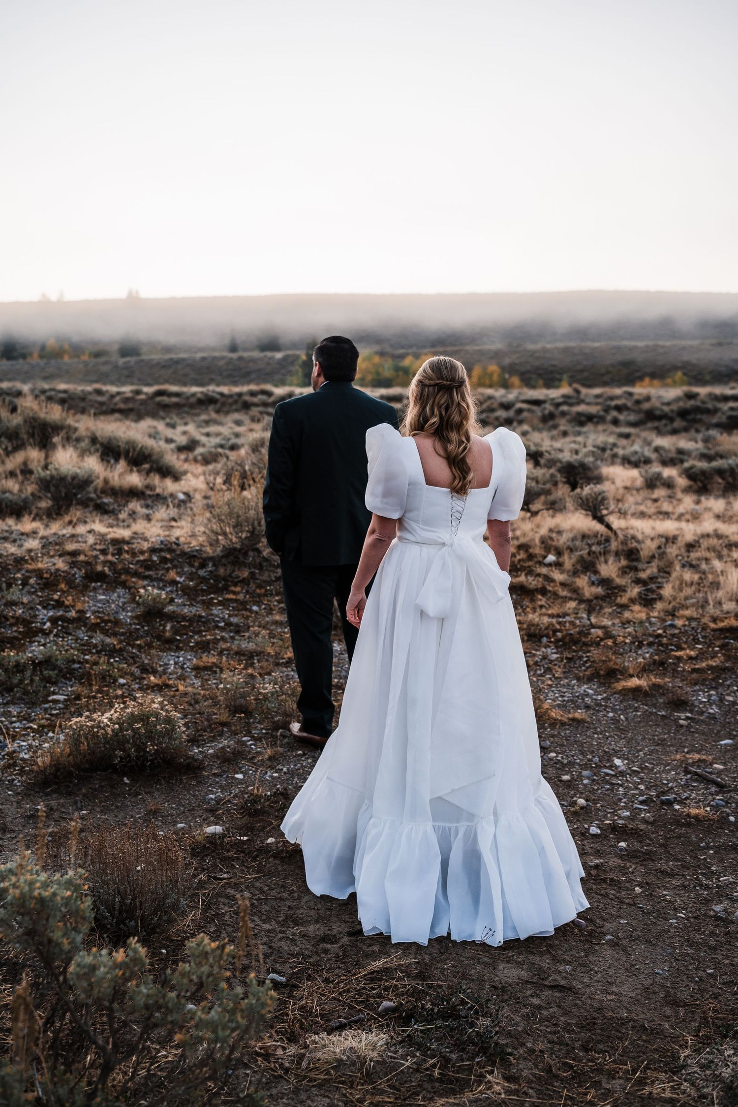 Three figures in white dresses stand together against a desert landscape at sunset.