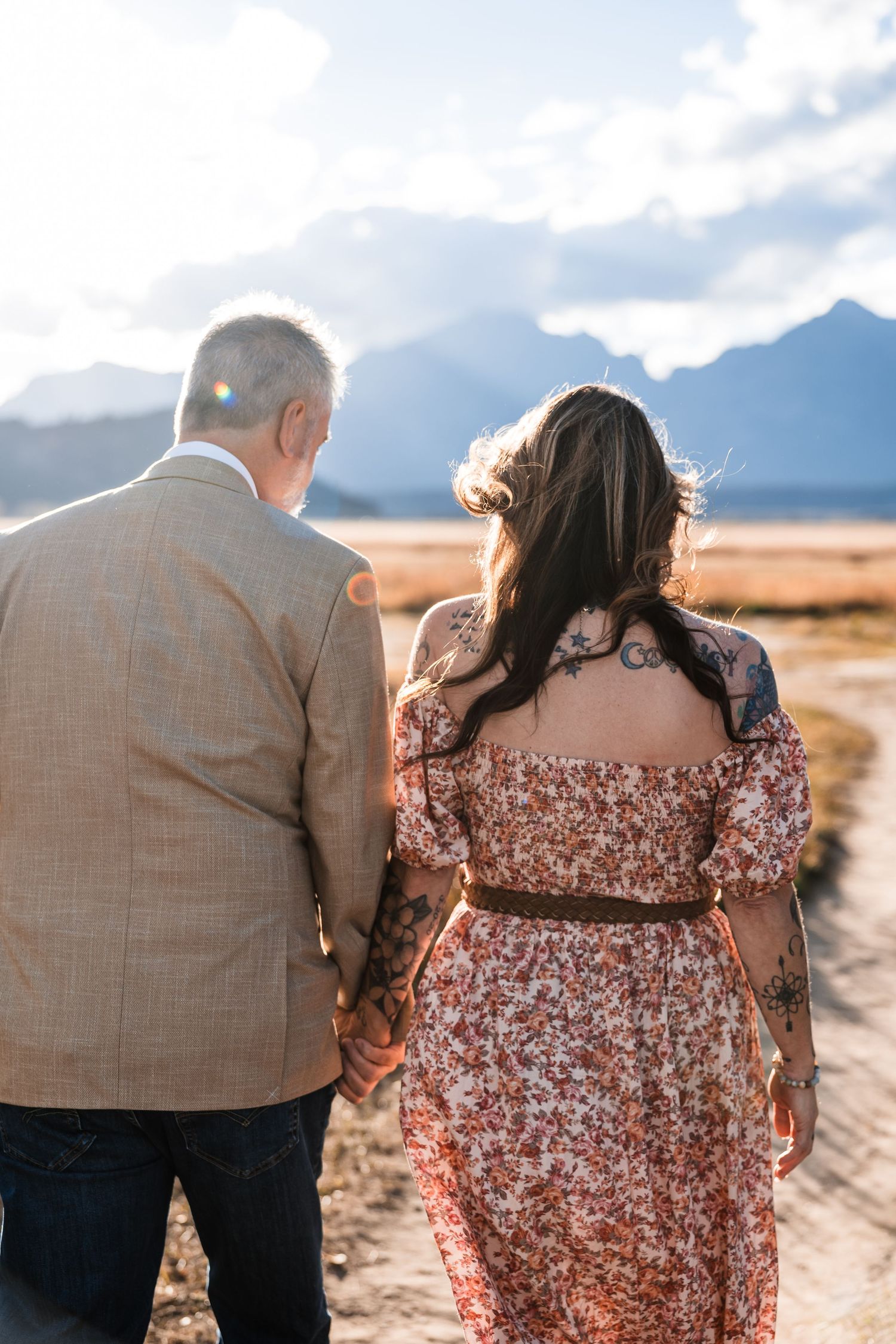 A couple in casual attire embraces while standing in an open field with mountain peaks in the distance.