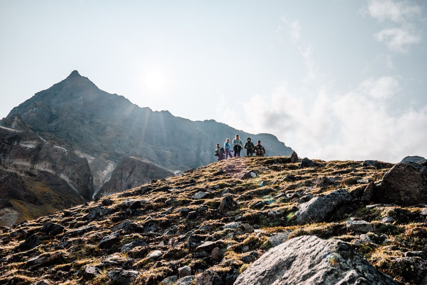 Hikers silhouetted on a rocky mountain ridge against a dramatic mountain peak and cloudy sky.