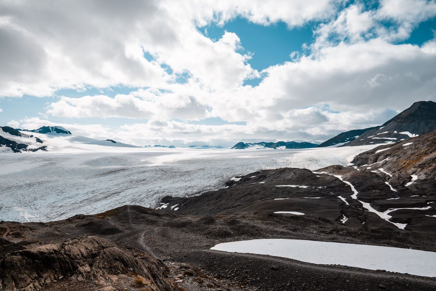 Dramatic mountain landscape with snow-covered glaciers and rocky terrain under a cloudy blue sky.