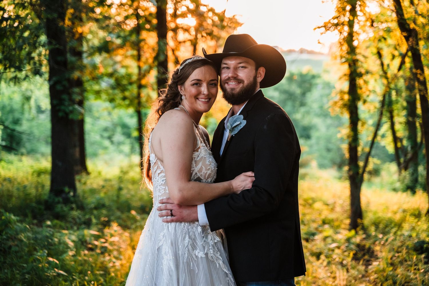 A couple shares an intimate moment in formal wedding attire against a sunset-lit forest backdrop.