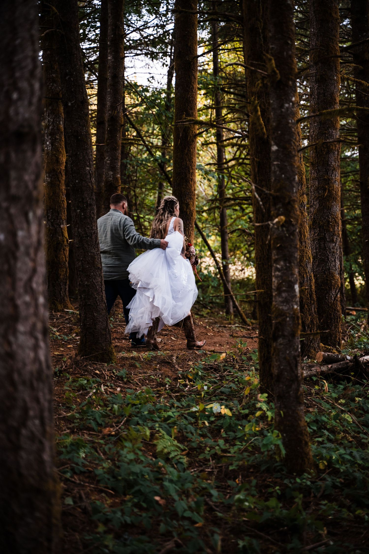 A couple walks through a forest path with the bride's white dress trailing behind.