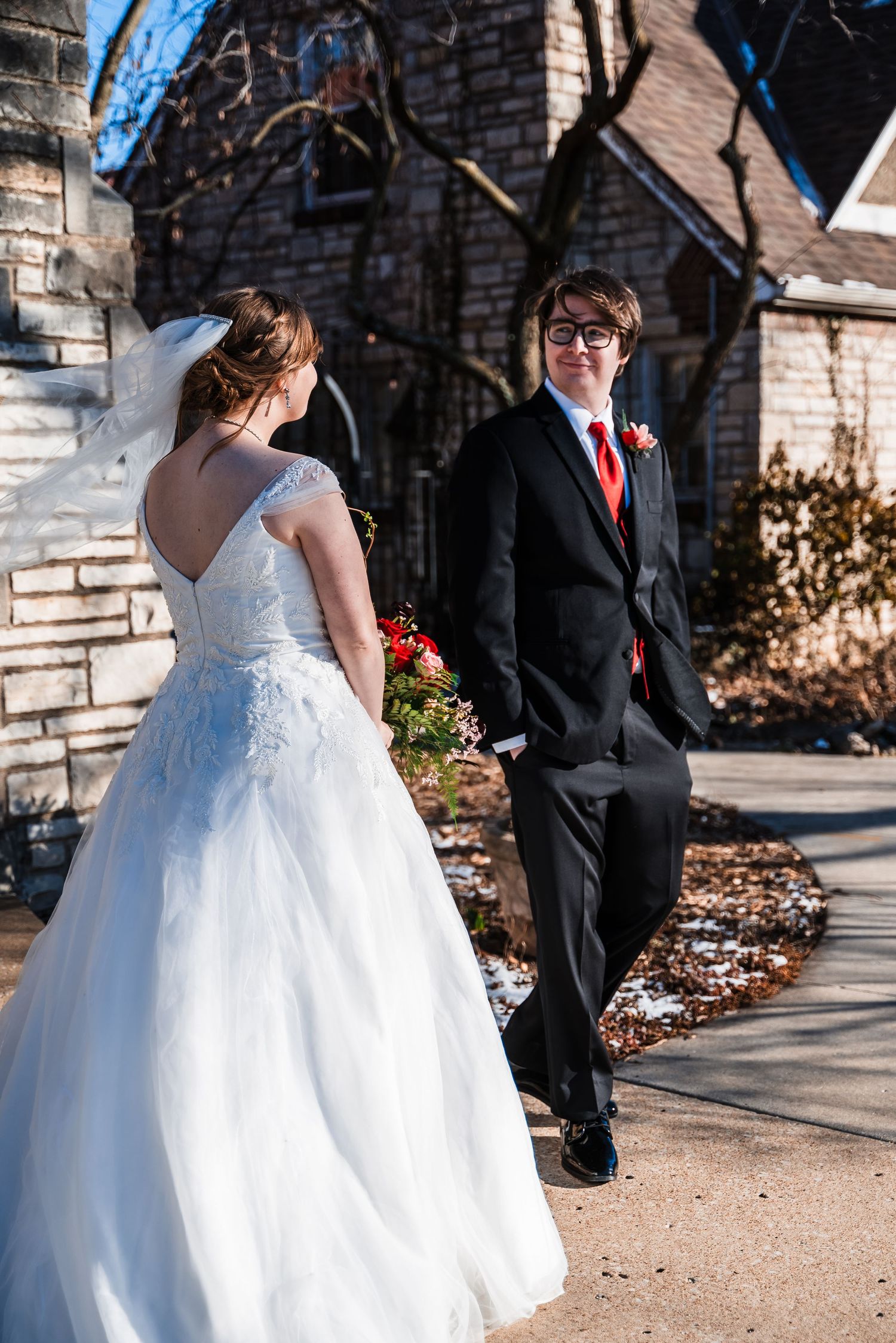 Bride in white gown and groom in black suit with red tie pose outside a stone church.
