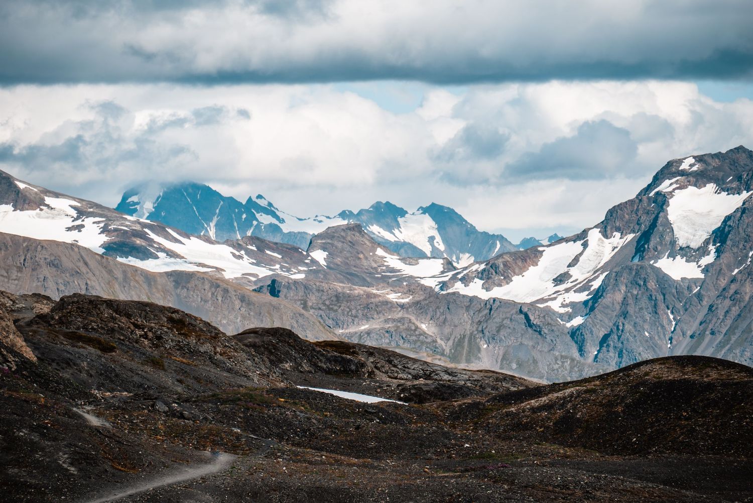 Rugged mountain landscape with snow-covered peaks and rocky terrain under cloudy skies.