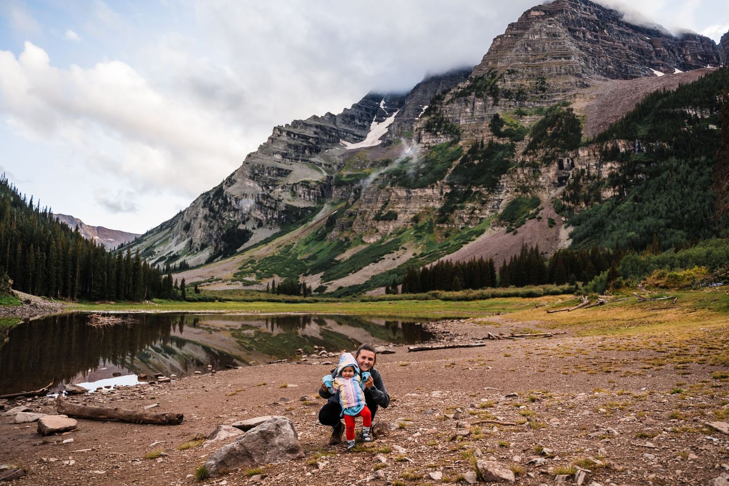 Two people explore the rocky shoreline of an alpine lake beneath towering mountain peaks.