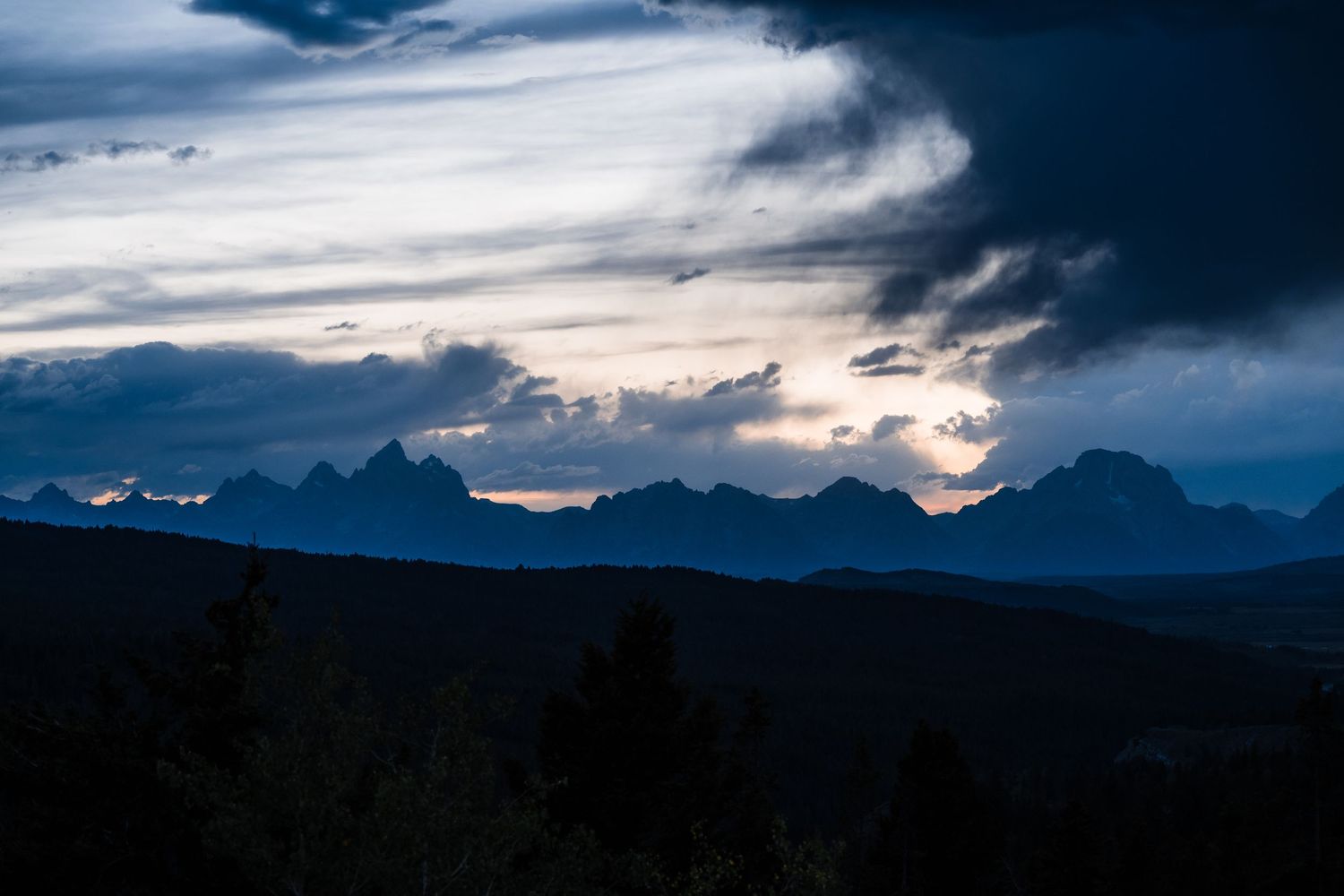 Dramatic sunset behind mountain silhouettes with dark moody clouds and golden light breaking through in wilderness landscape.