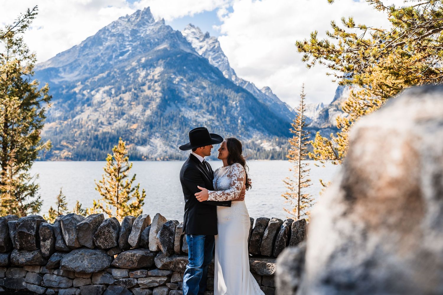 Newlyweds embrace against stunning mountain backdrop with snow-capped peaks and evergreen trees.
