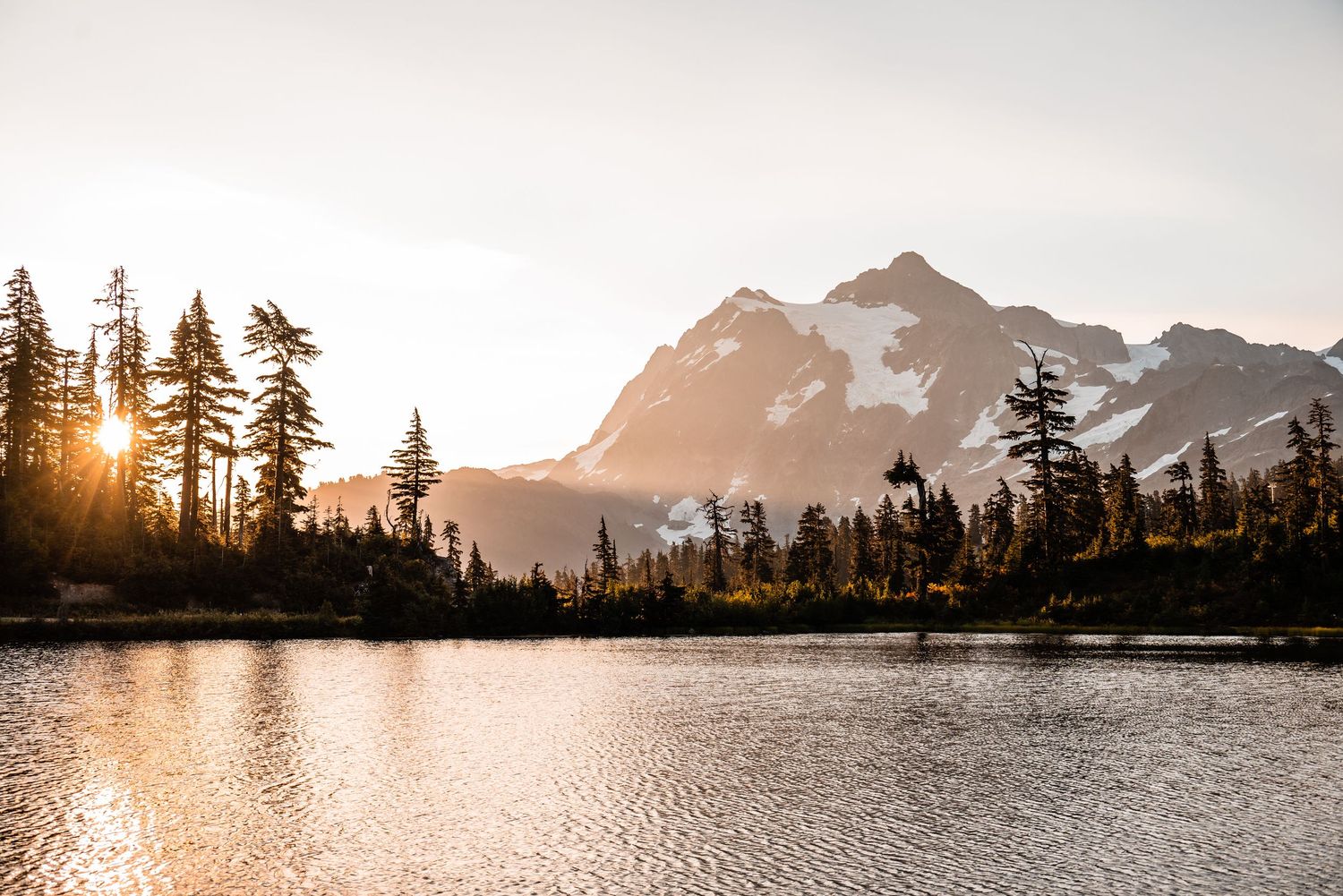 Golden sunset illuminates a serene mountain lake surrounded by evergreen trees and rugged peaks.