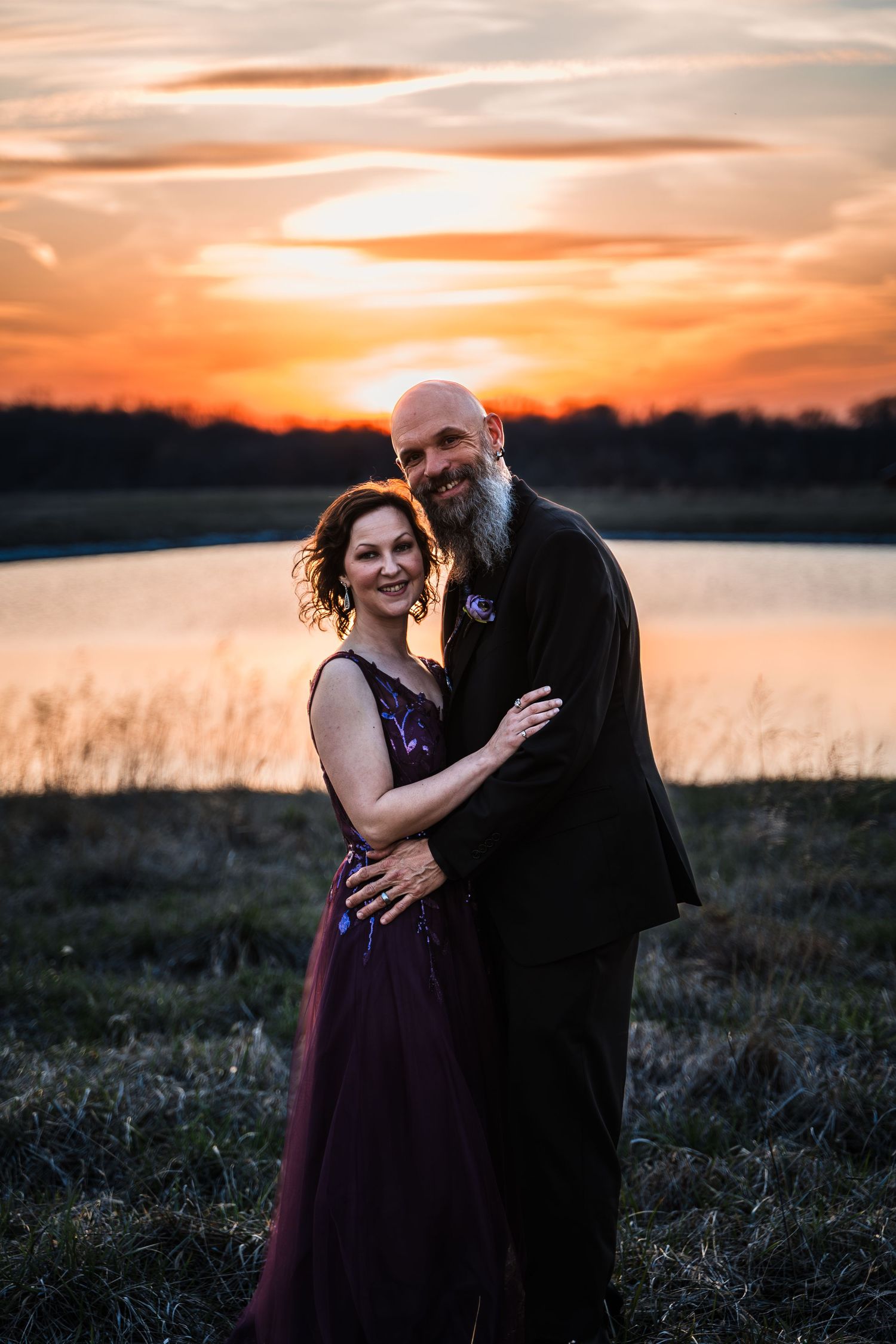 Wedding couple embracing at golden sunset near reflective pond, bride in burgundy gown.