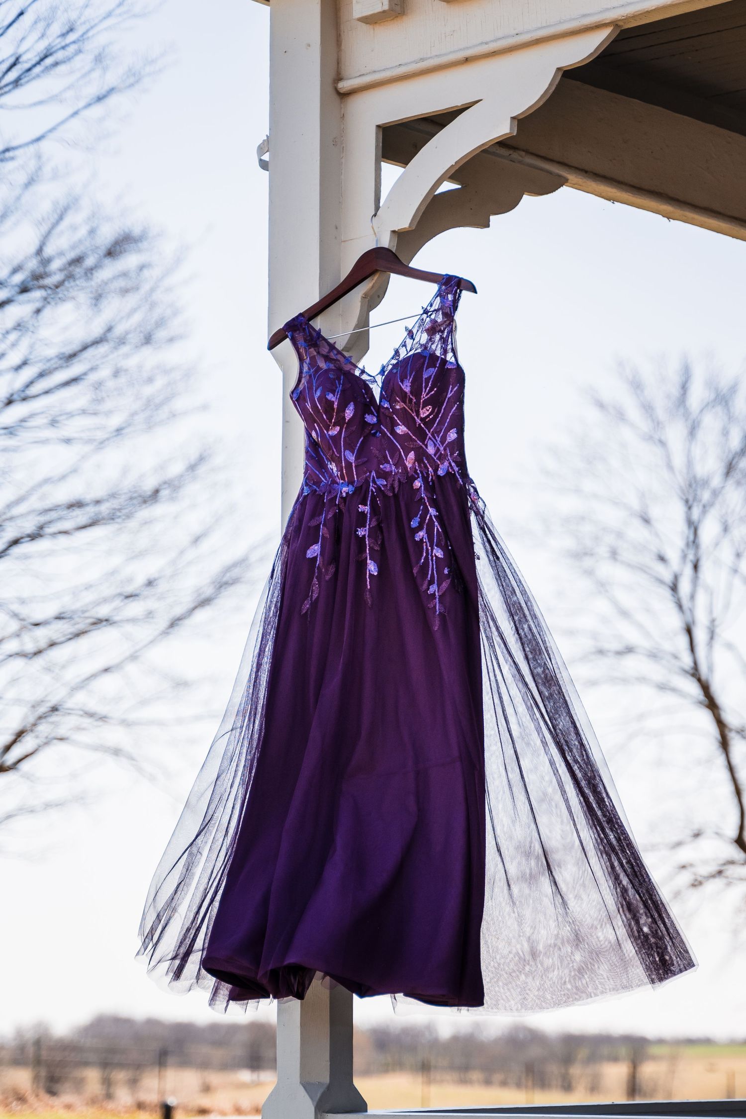 Purple embroidered tulle cocktail dress hanging on a white gazebo post outdoors in winter.