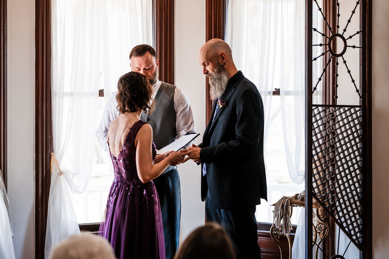 A couple exchanges wedding vows with an officiant indoors, bride in purple dress, groom in suit.