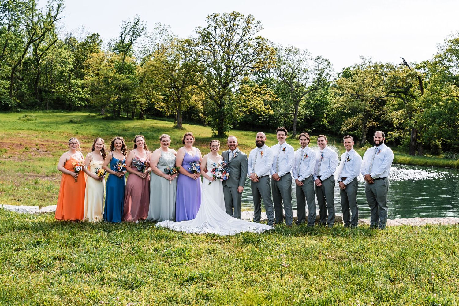 Wedding party poses by a pond in lush green landscape, bridesmaids in colorful dresses with groomsmen in gray suits.