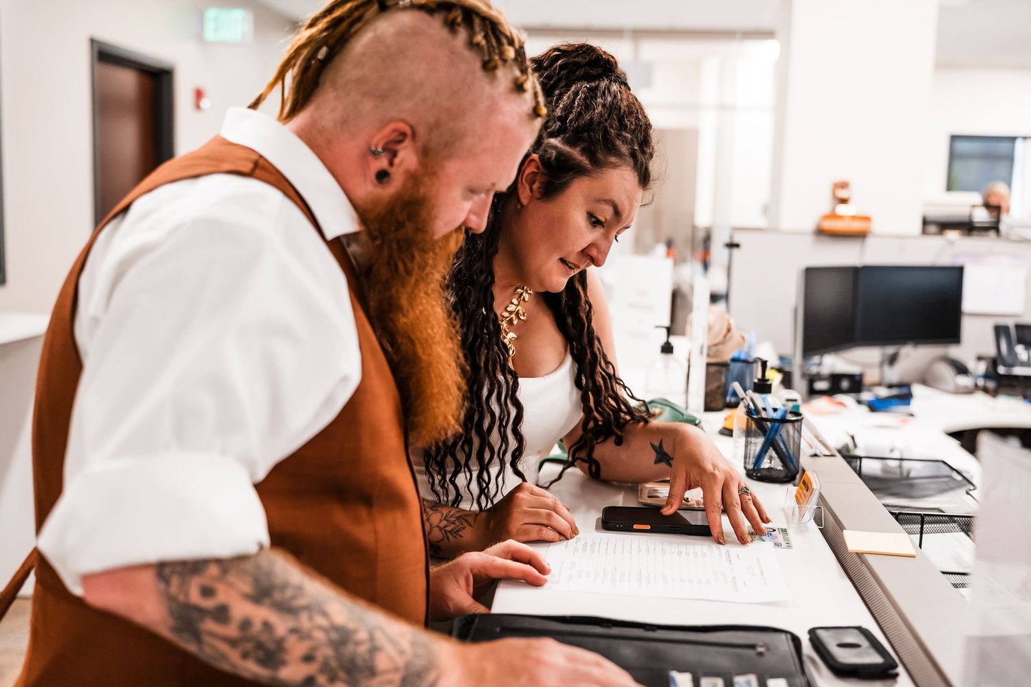 Tattooed bride and groom signing marriage license documents together at courthouse registration desk.