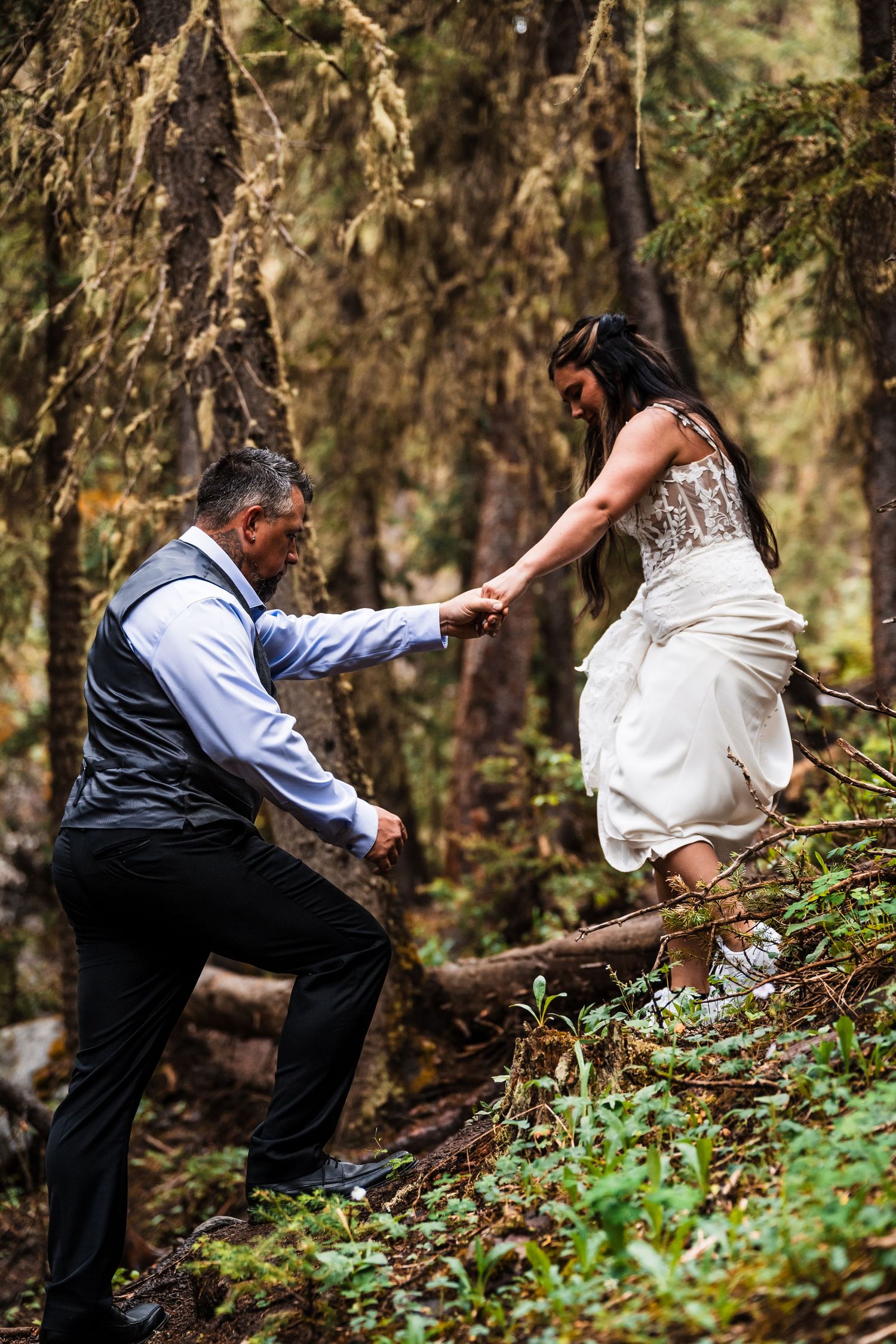 Groom helps bride navigate forest terrain during adventurous outdoor elopement in the woods.