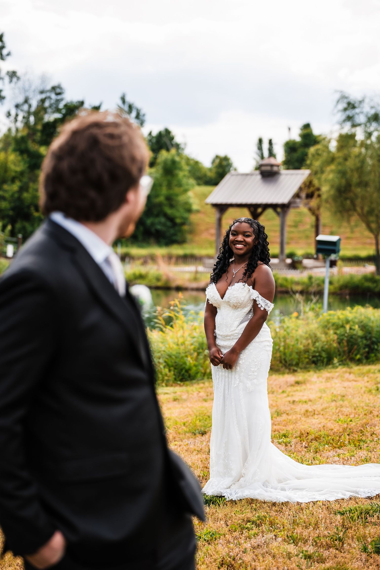 Bride in white off-shoulder gown smiles at groom during first look at outdoor wedding venue with gazebo.