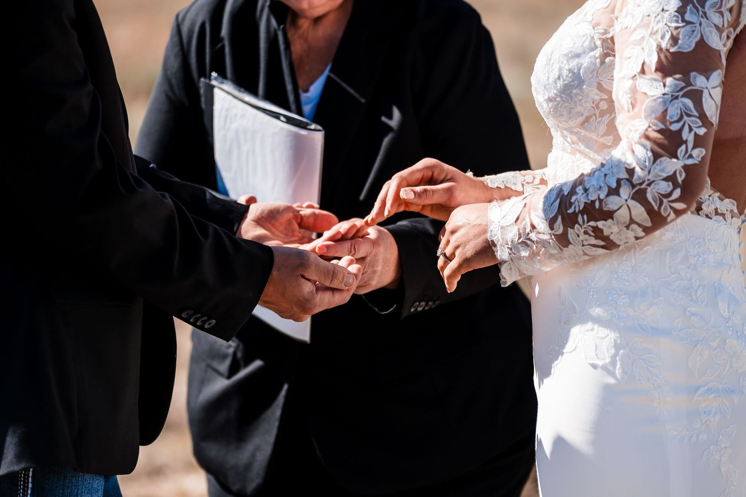 Close-up of hands exchanging wedding rings during an outdoor ceremony with lace dress details visible.