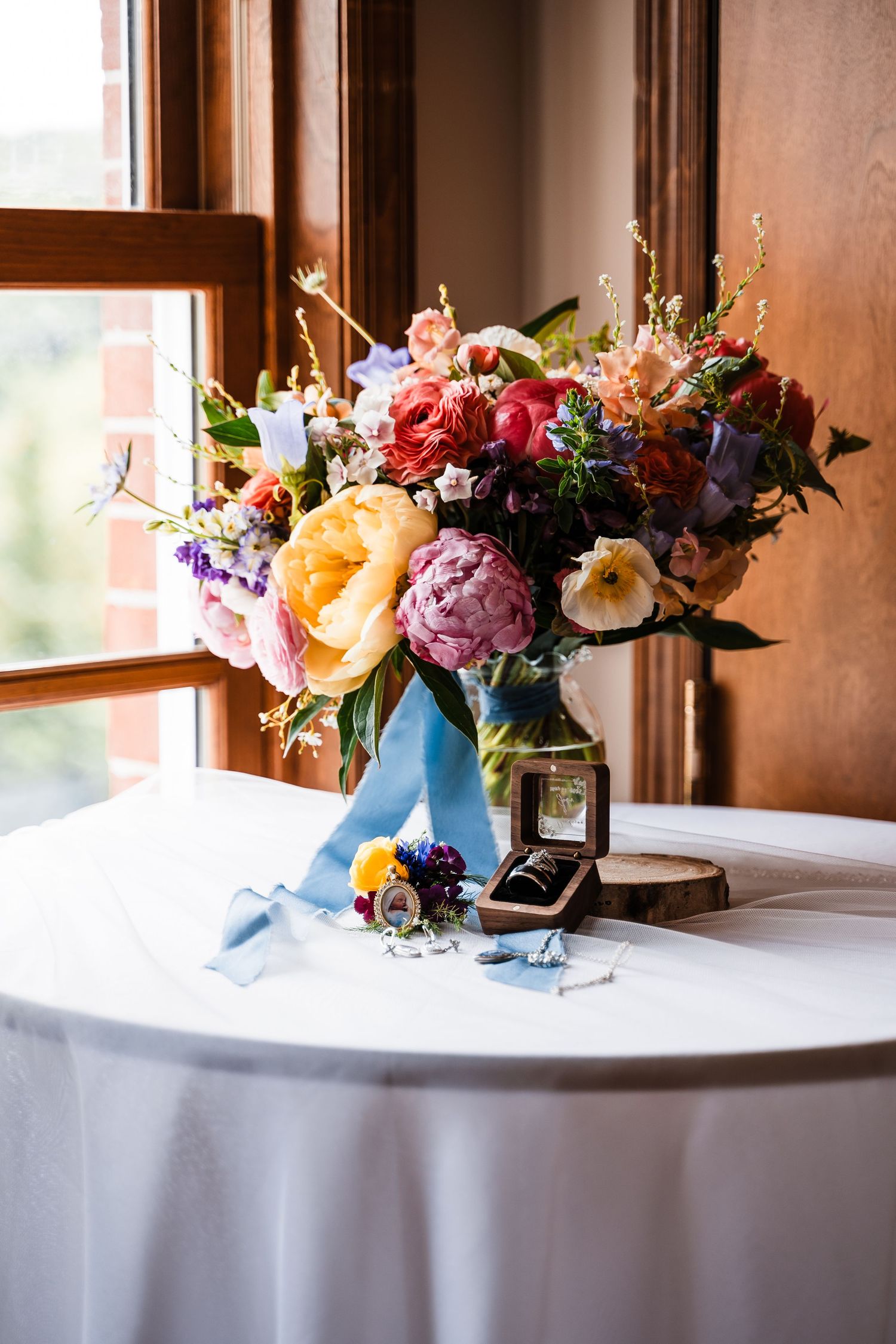 Vibrant floral arrangement with peonies, roses and mixed flowers in light blue vase on white tablecloth by window.