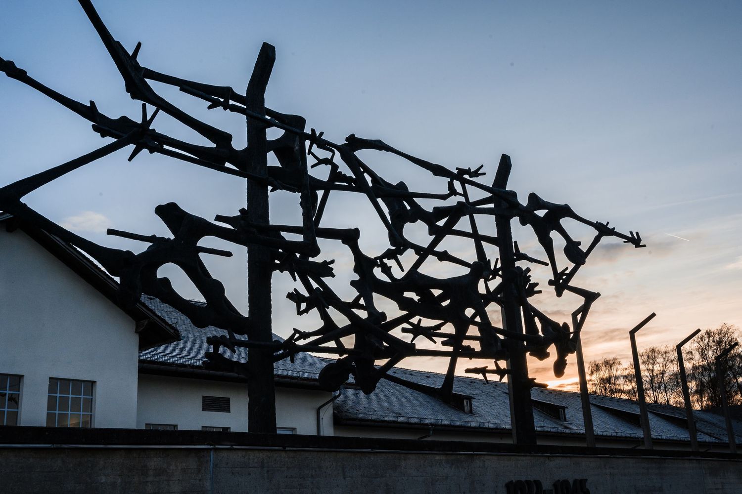 Silhouette of barbed wire fencing against a dusky sky at a historical concentration camp memorial site.