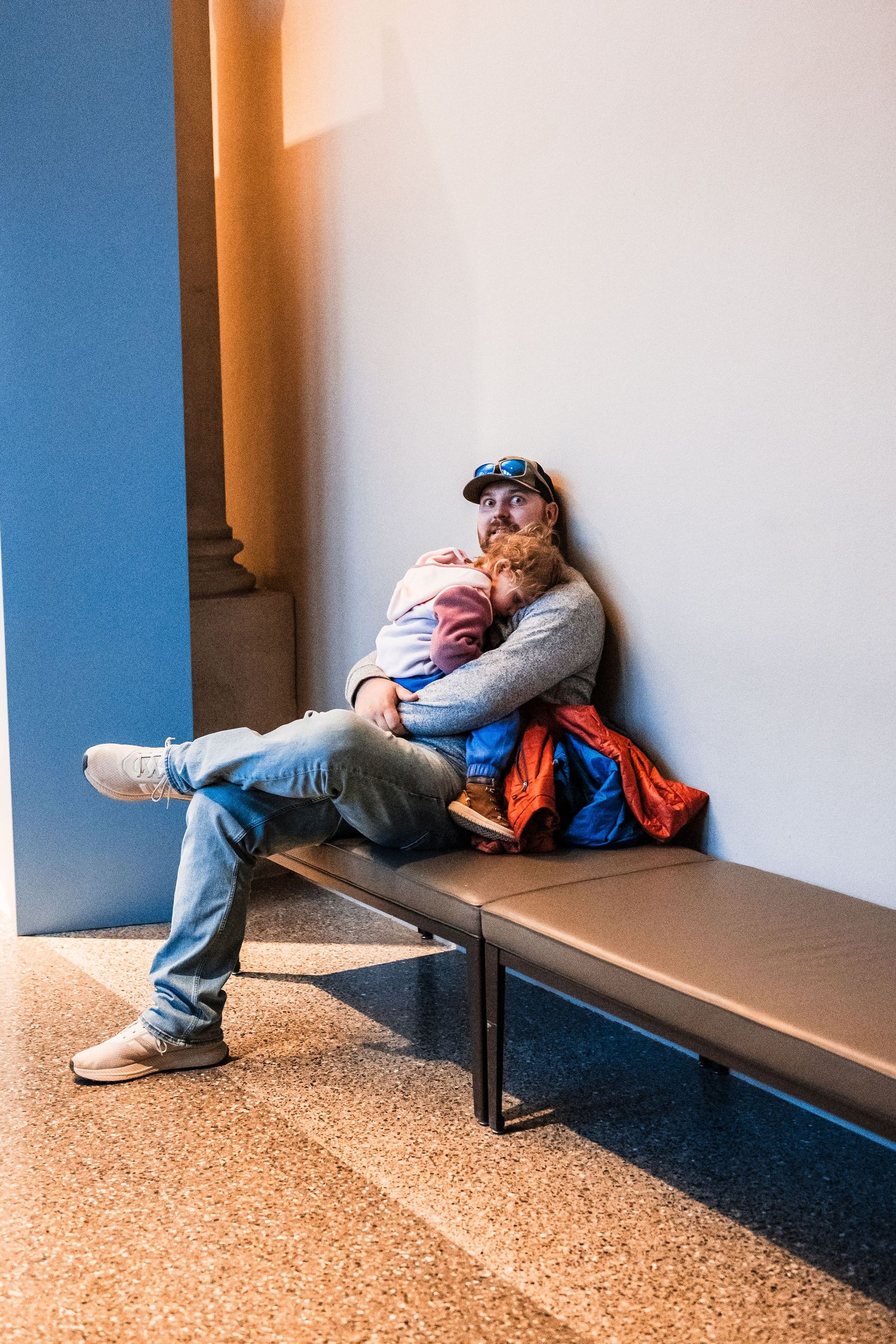 Person lying down on bench in museum hallway with orange wall and speckled floor.
