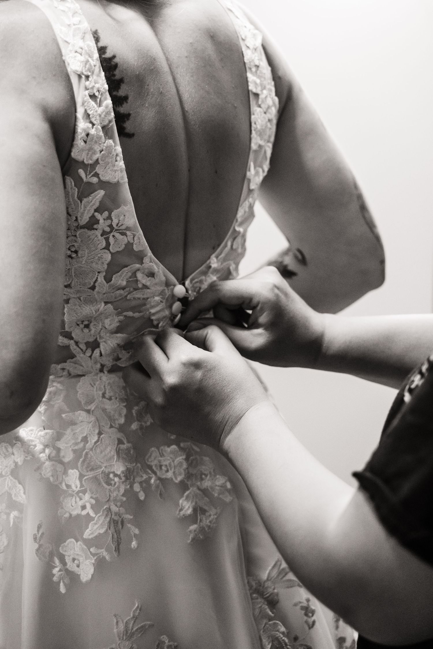 Hands fastening delicate lace buttons on the back of an elegant white floral wedding dress in black and white photography.