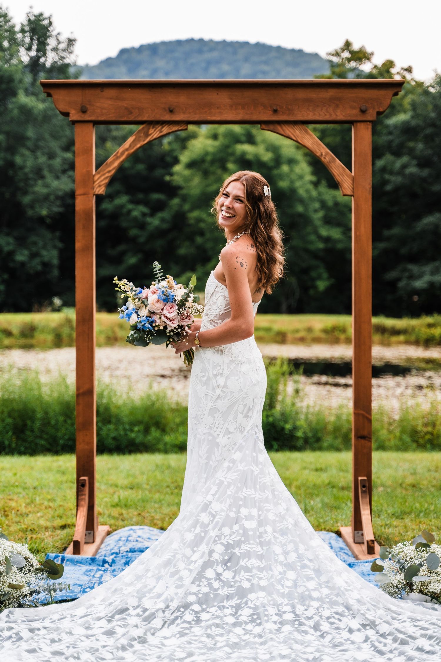 A white lace wedding dress with a long train displayed under a wooden arch in an outdoor garden setting.