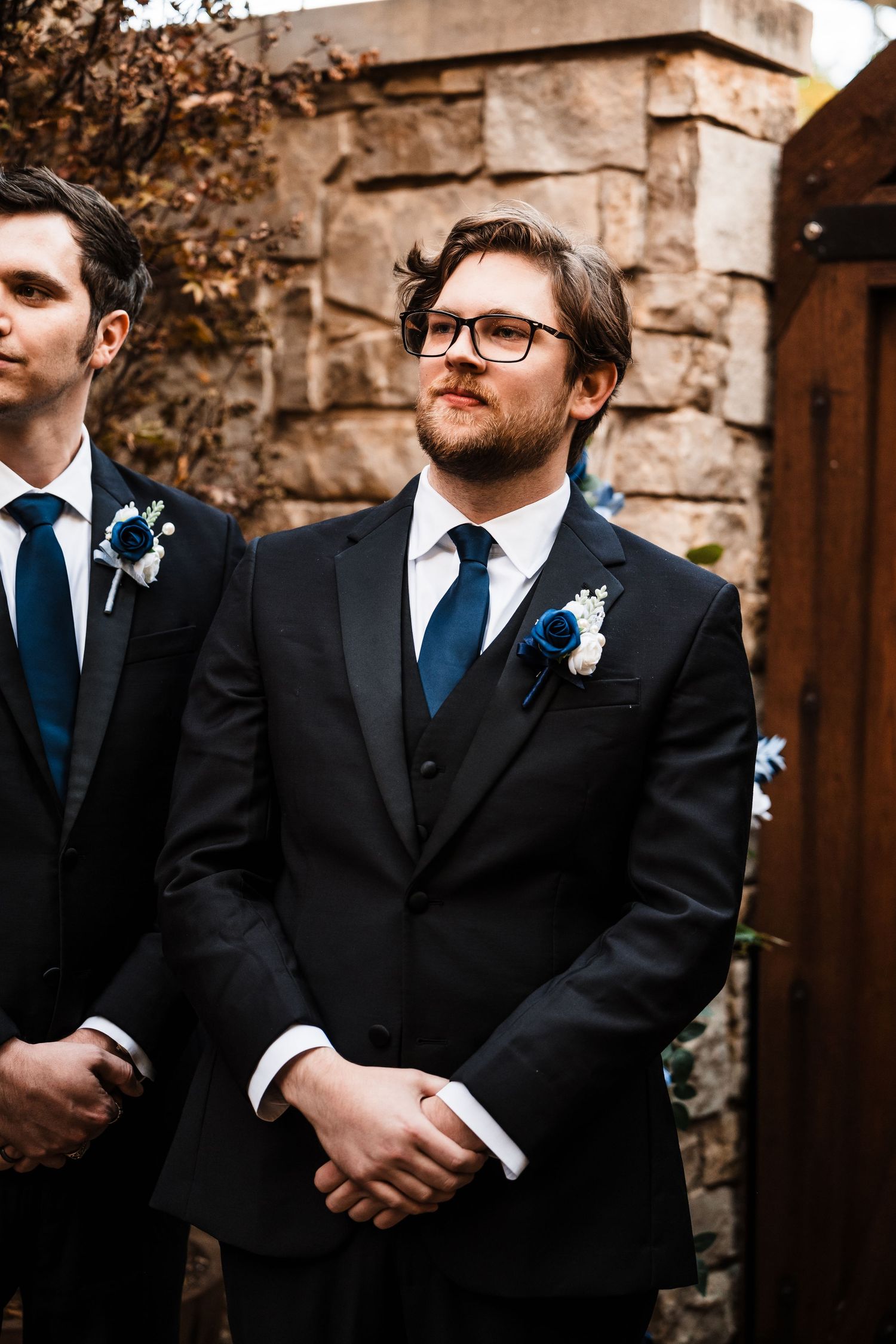 Two men in formal black suits and ties stand in front of stone architecture at outdoor event.