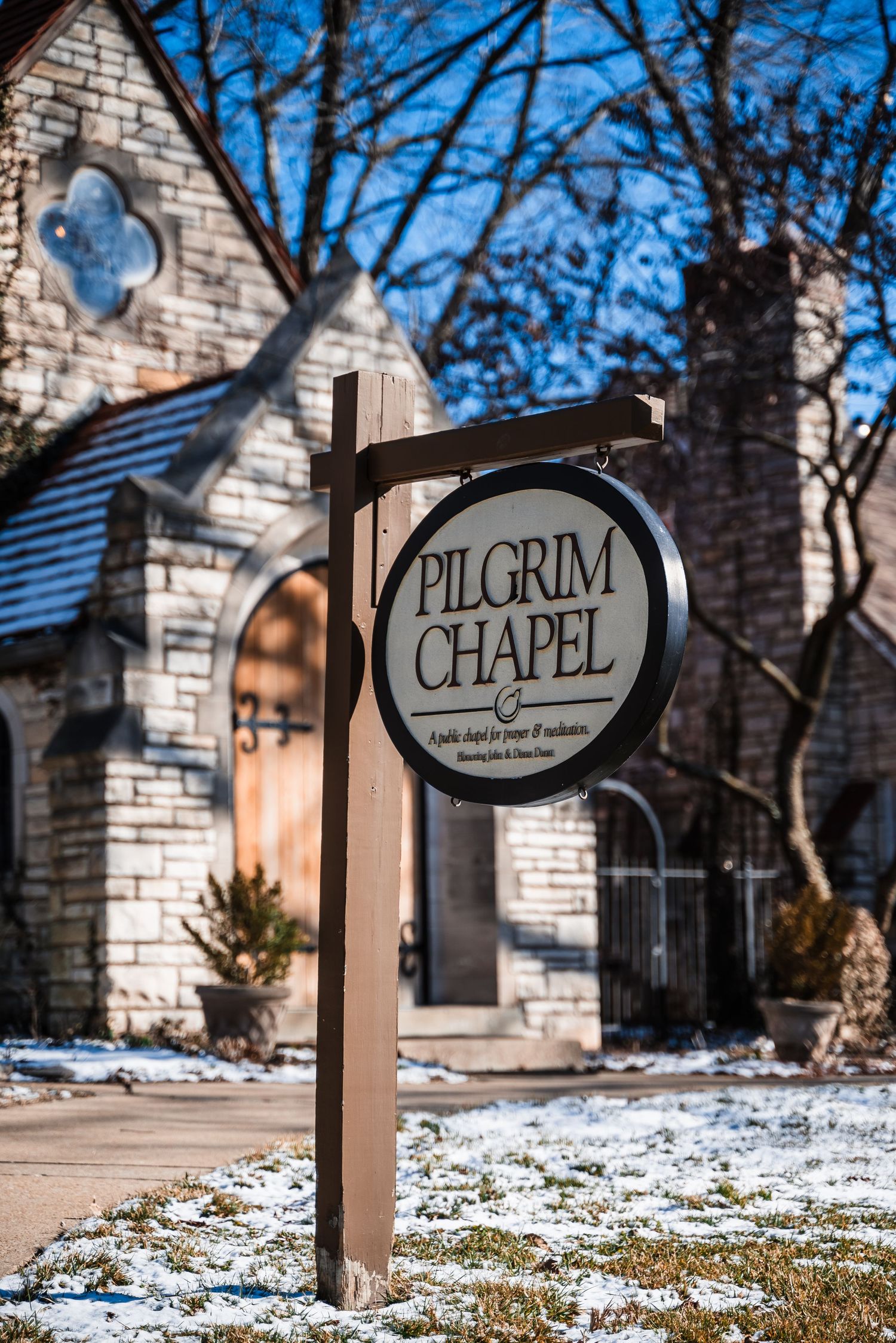 A rustic stone Pilgrim Chapel sign stands in front of a quaint church building on a snowy winter day.