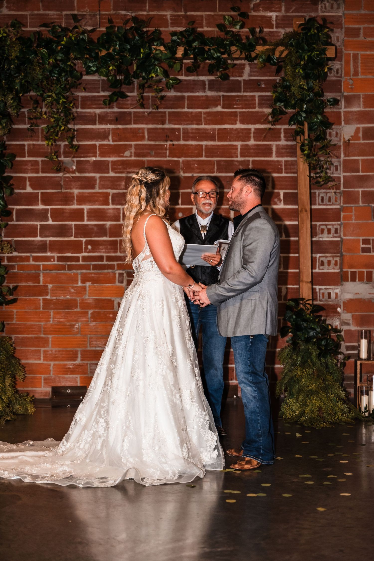Wedding ceremony in progress under a greenery arch against industrial brick wall backdrop.