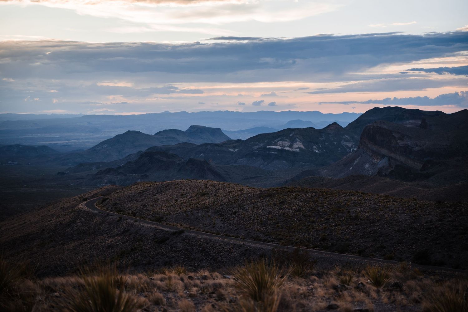 A dramatic mountain landscape at dusk with layered peaks stretching into the distance under a cloudy sky.
