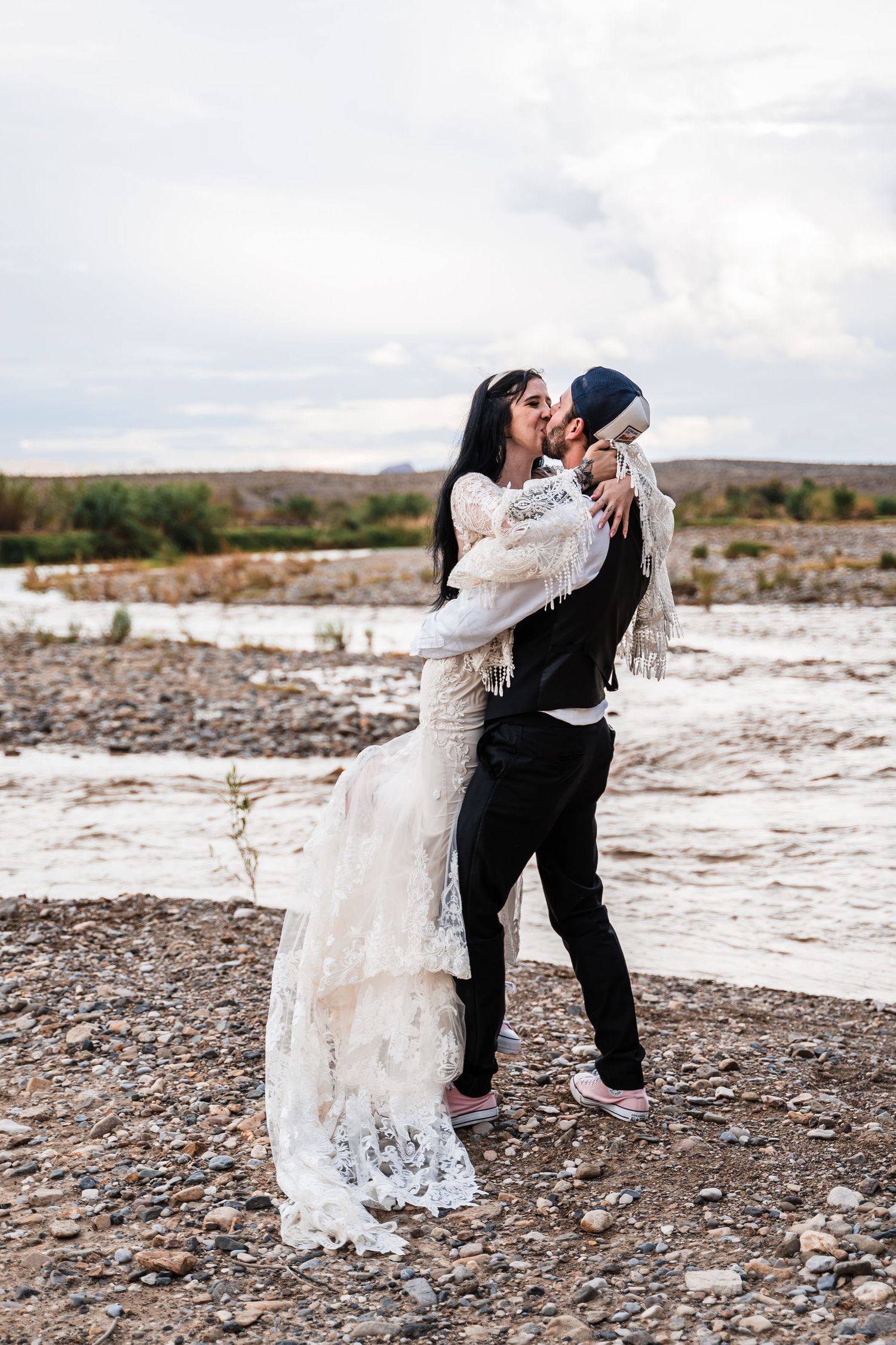 A couple in wedding attire embraces by a river with rocky mountains in the background.