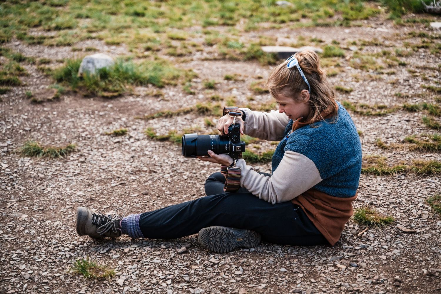 A photographer sits on desert ground while adjusting settings on a professional DSLR camera.