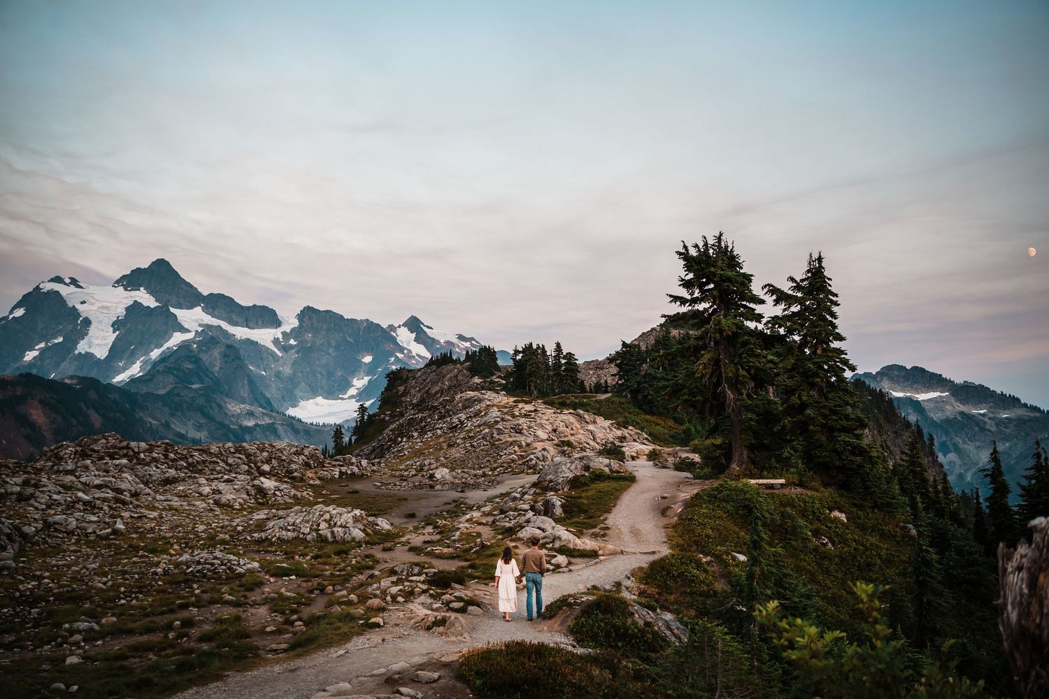 Snow-capped mountain peaks rise above a winding hiking trail through rugged alpine terrain at sunset.