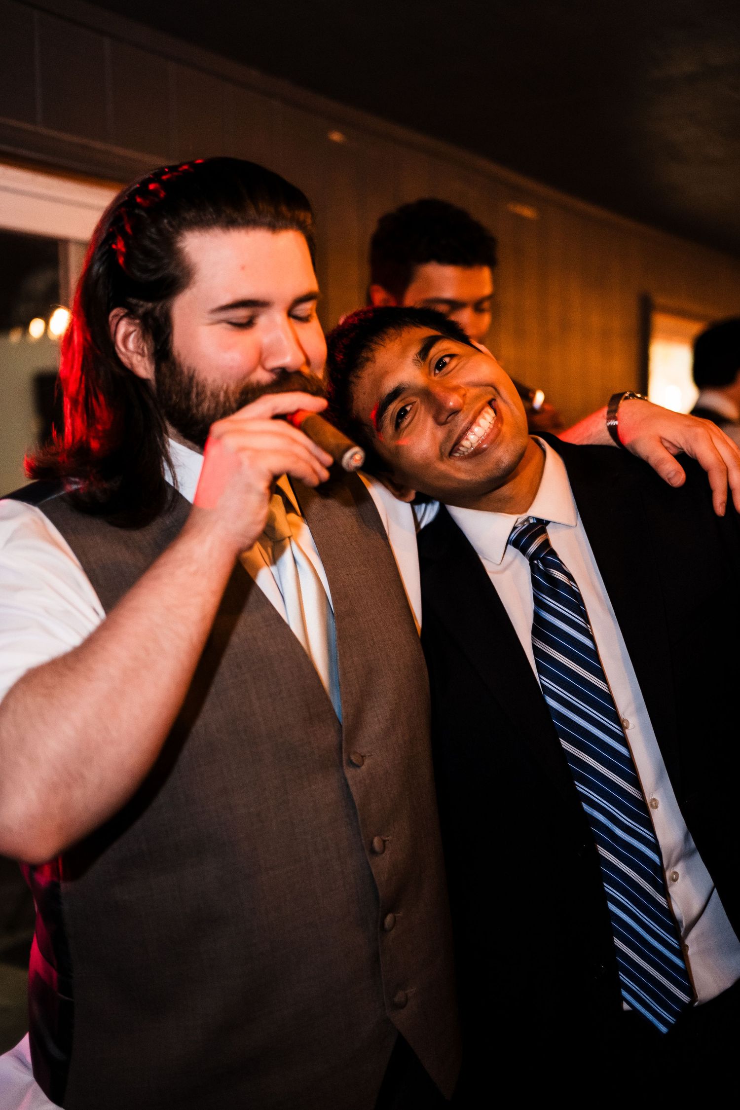 Two friends celebrate joyfully at a wedding reception with one wearing a vest and another in a suit and tie.