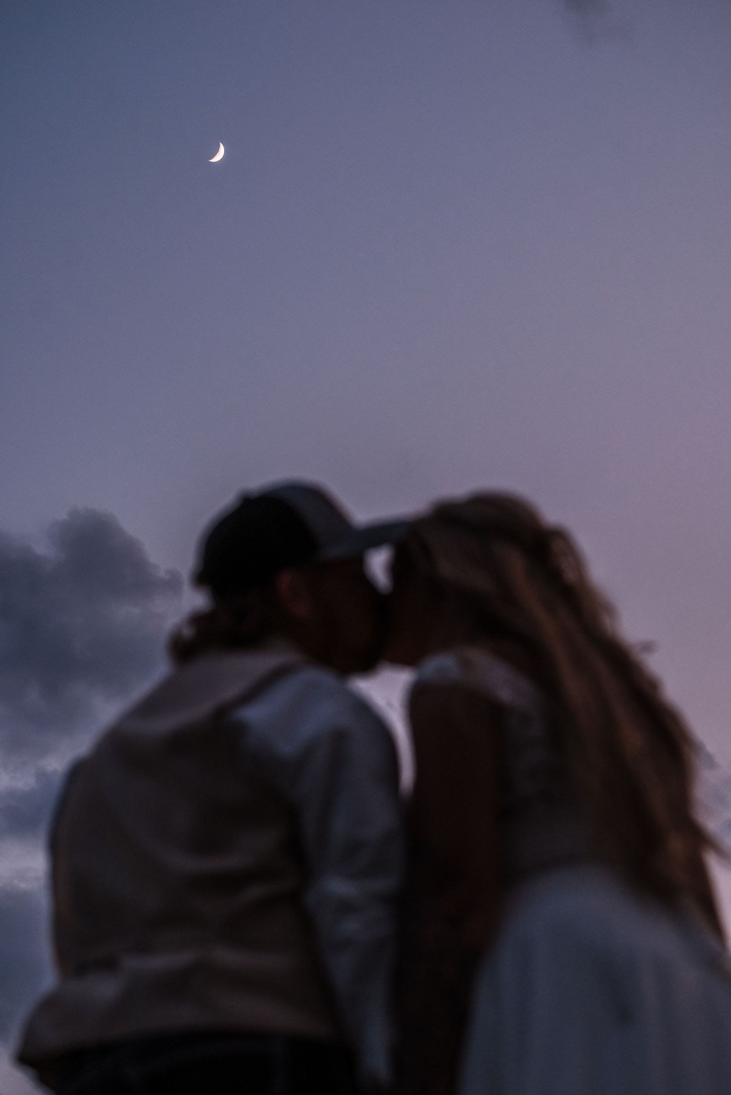 A romantic silhouette of a couple kissing under a crescent moon at dusk.