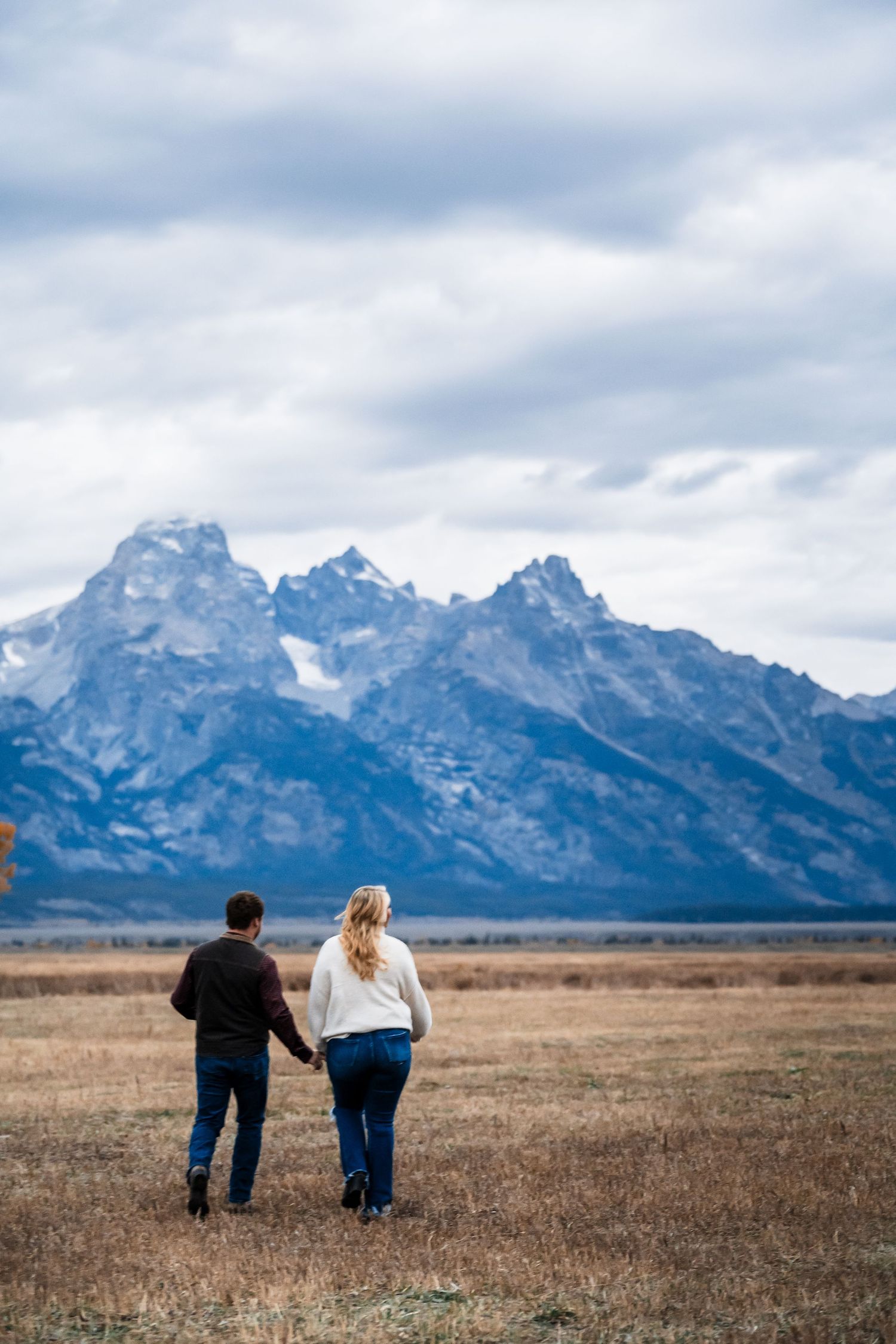 A couple walks hand in hand across a vast plain with the majestic Teton mountain range rising dramatically in the background.