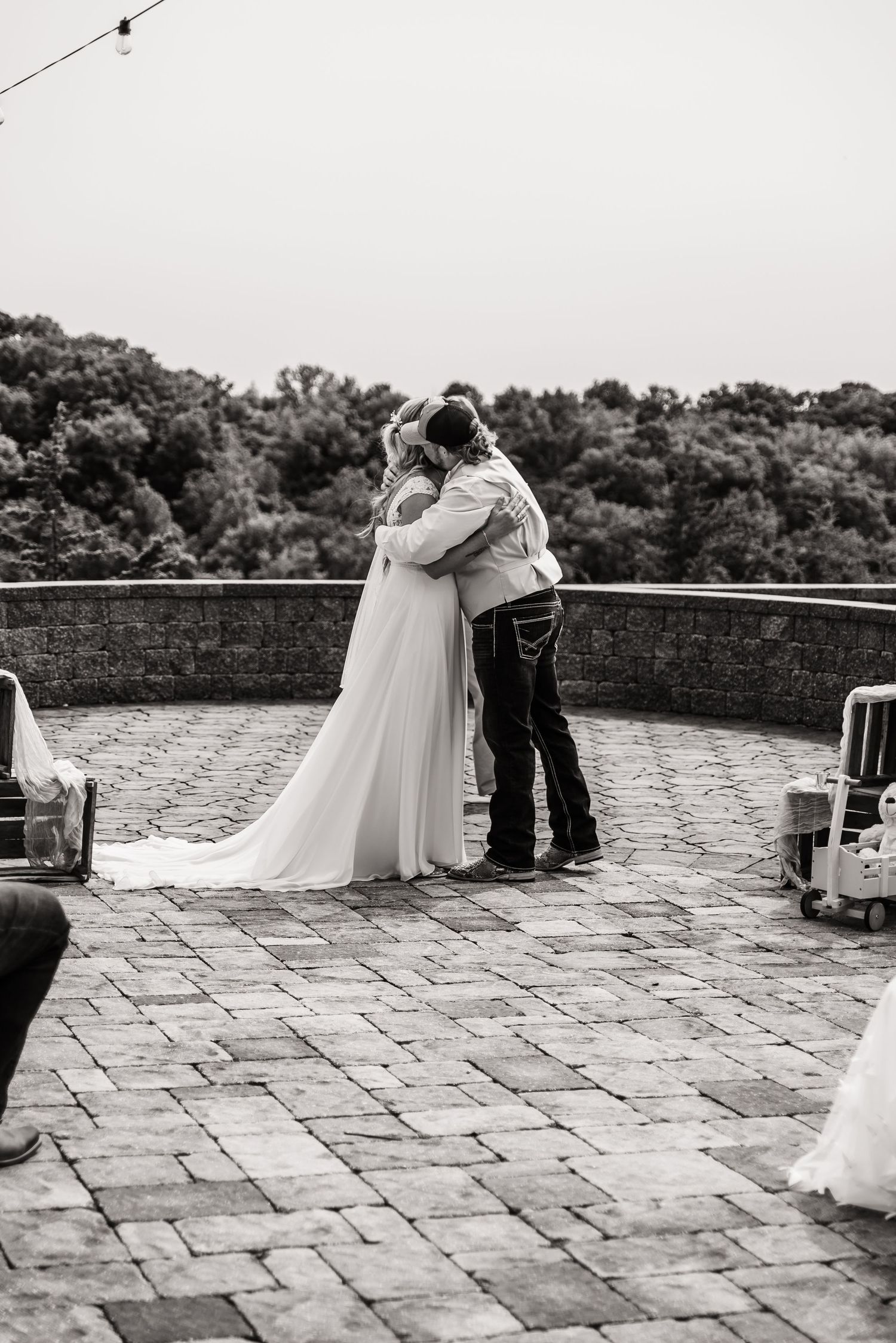 A black and white wedding photo showing an intimate embrace on a stone patio surrounded by trees.