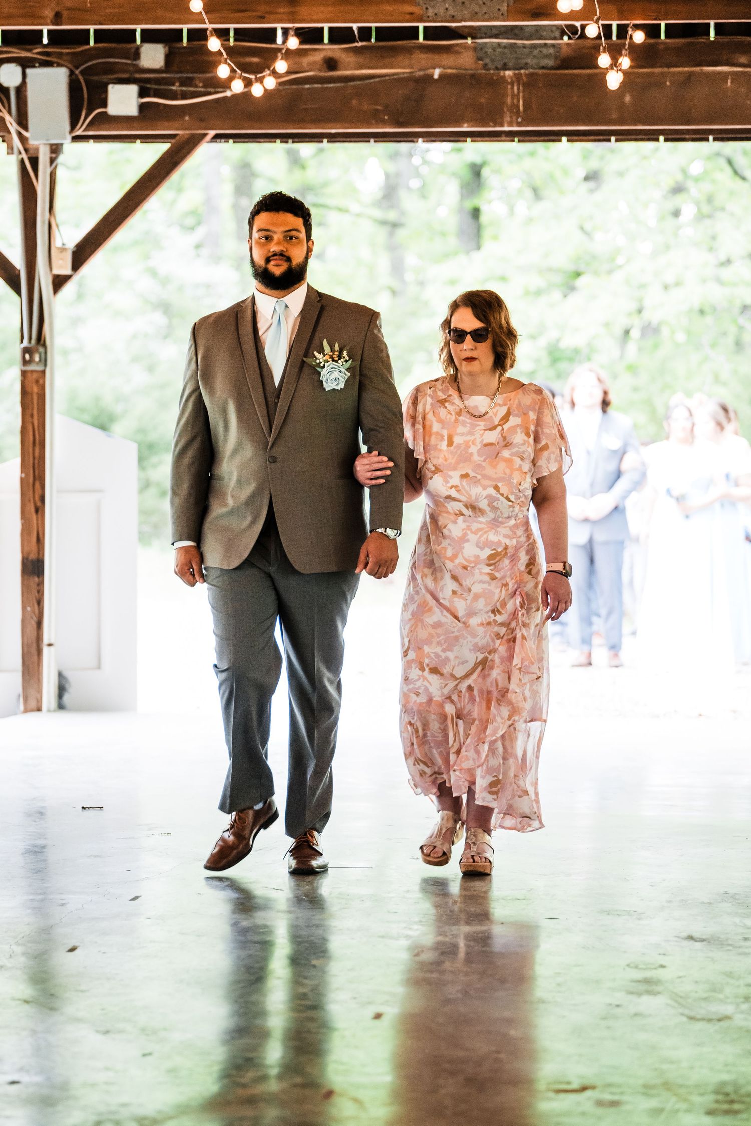 A couple walks down an aisle under a wooden pavilion adorned with string lights.