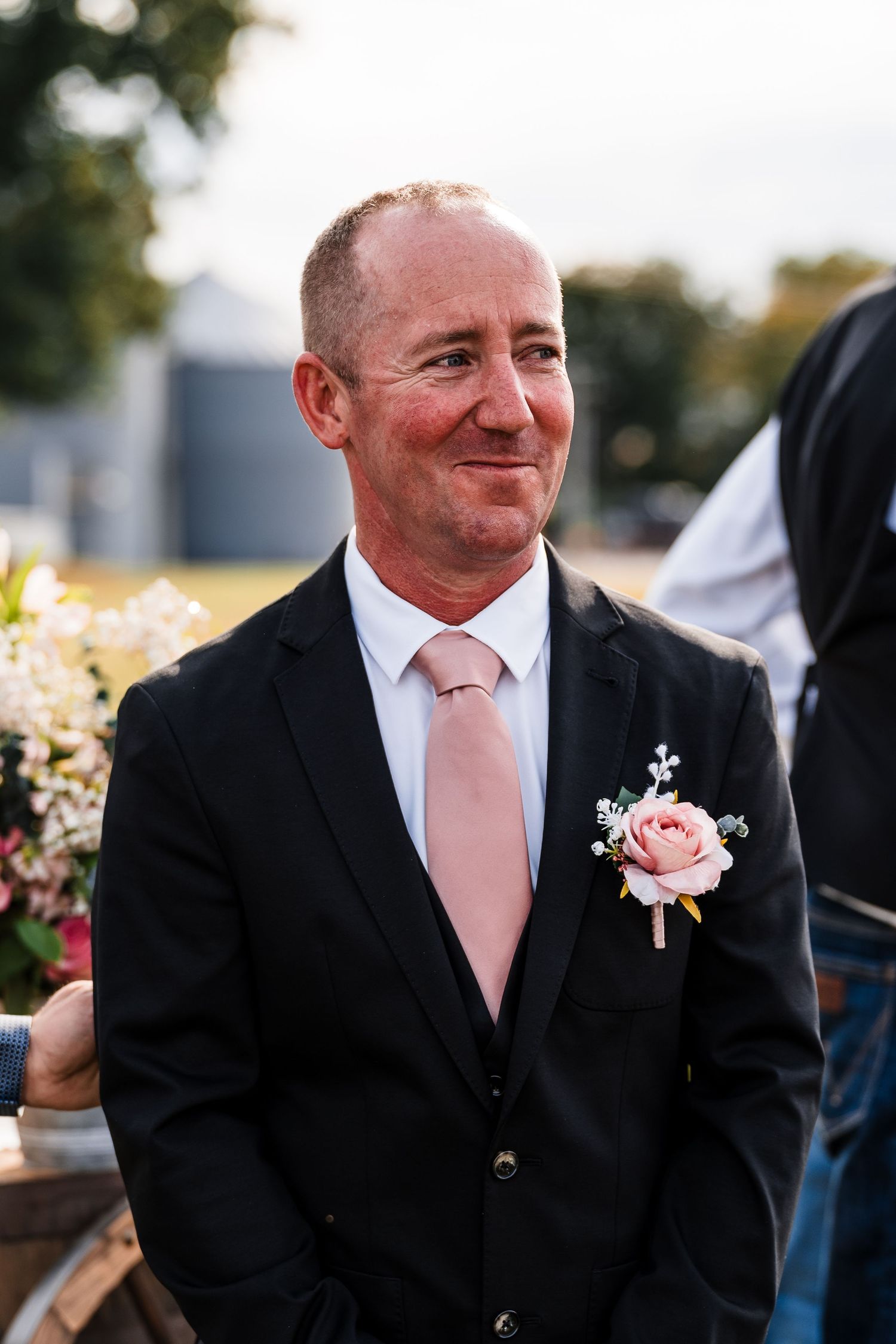 A groom in a black suit with pink tie and rose boutonniere smiles during an outdoor wedding.