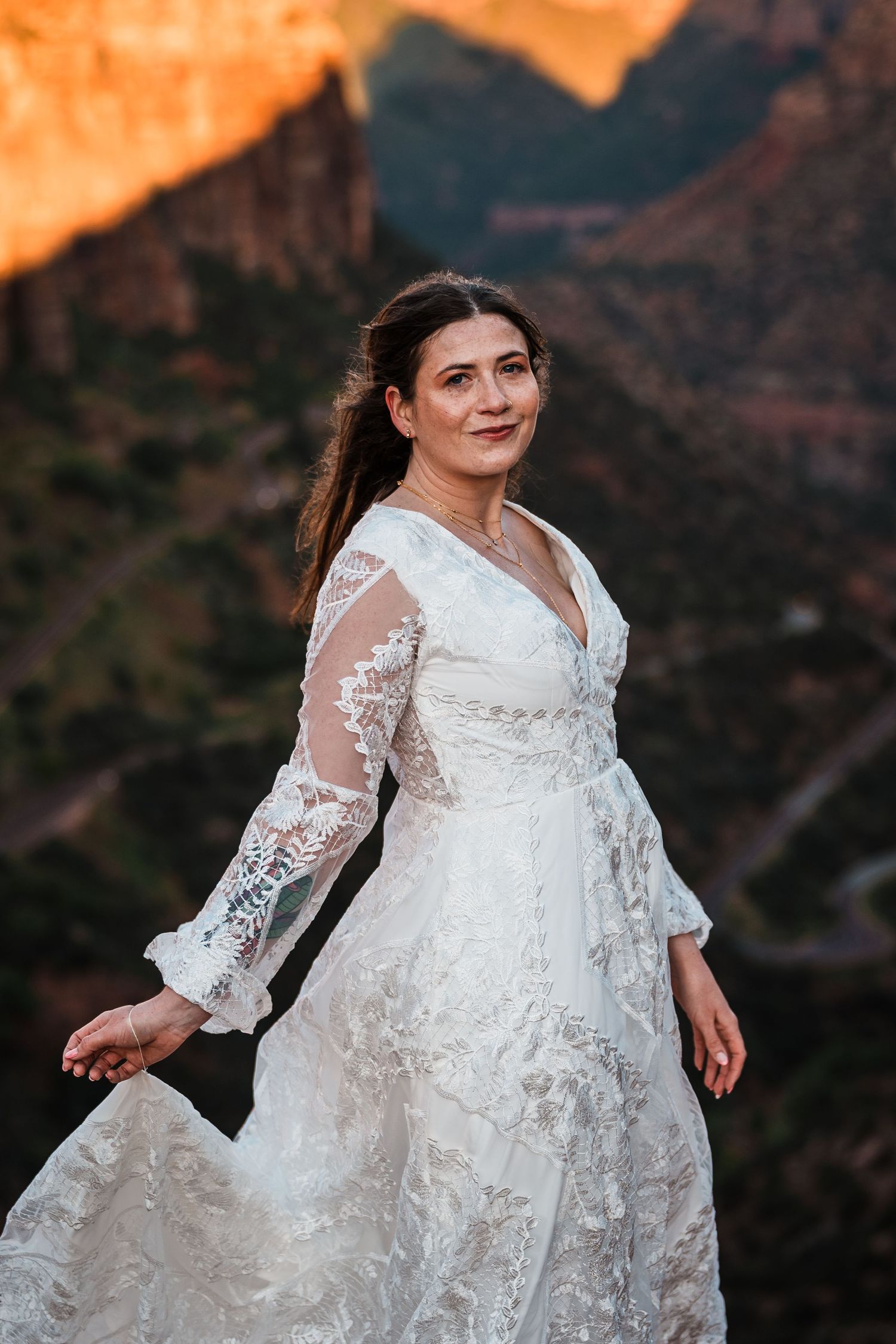 Bride in lace long-sleeve wedding dress poses at sunset with red canyon mountains behind her.