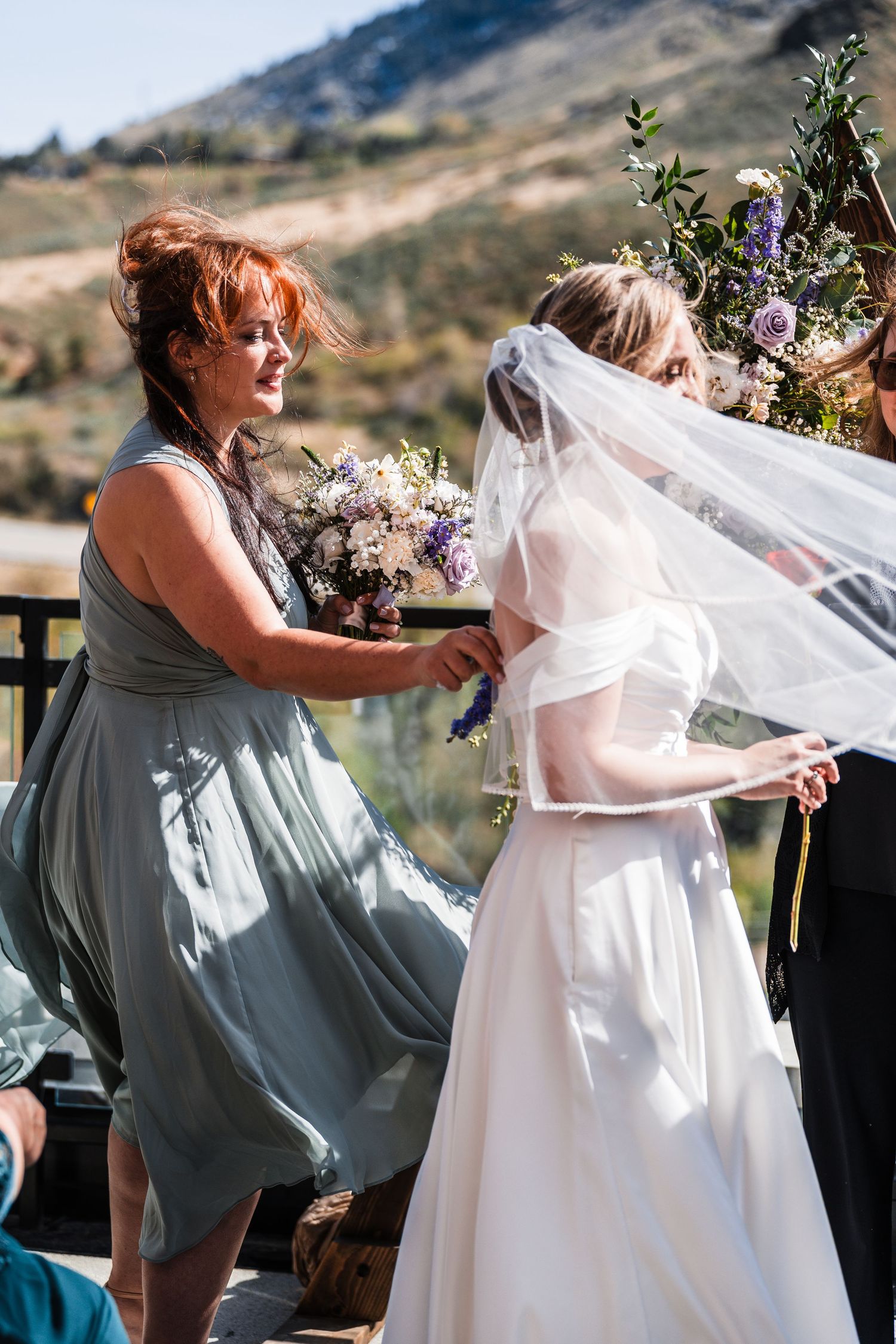 Bridesmaid in sage green dress holding bouquet and adjusting brides veil outdoors at mountain wedding ceremony.