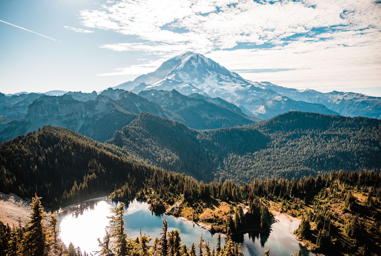 Panoramic mountain landscape with snow-capped peaks, evergreen forests and alpine lakes under cloudy skies.
