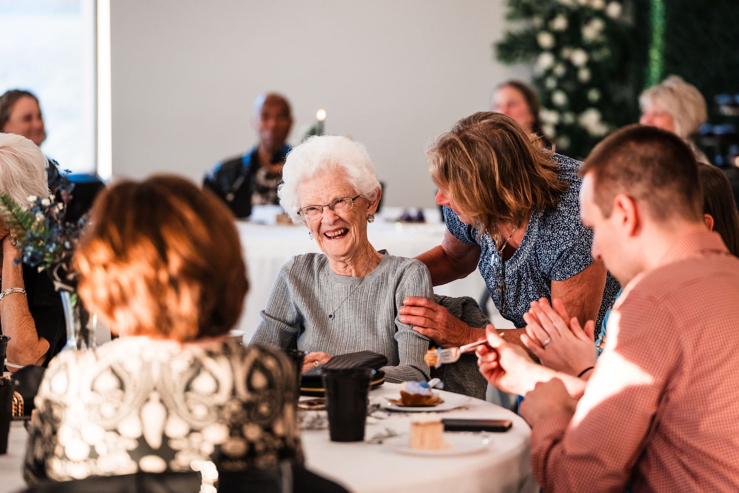Elderly woman laughing joyfully with guests at wedding reception dinner table in bright modern venue.